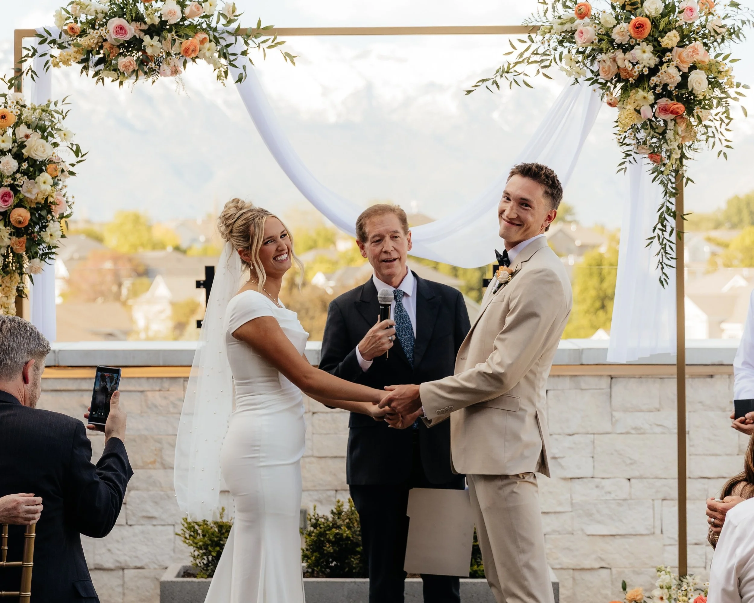 Bride and groom stand hand in hand in front of the officiator smiling at the audience.