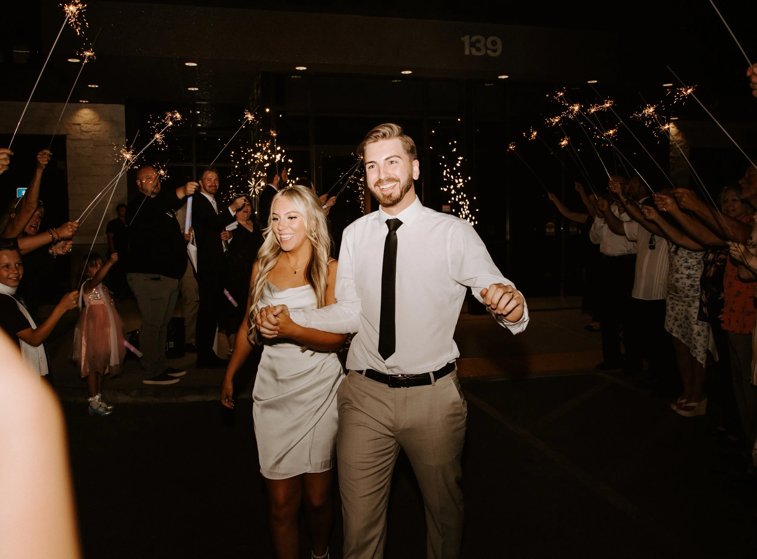Bride and groom walk holding hands with sparklers held by guests in the background.