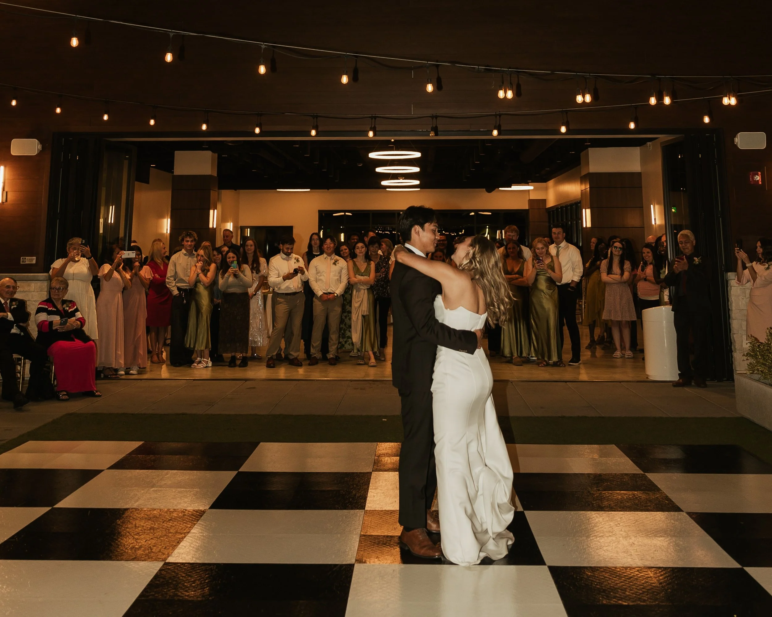 Bride and groom are dancing on black and white tile flooring while their guests watch and take pictures.