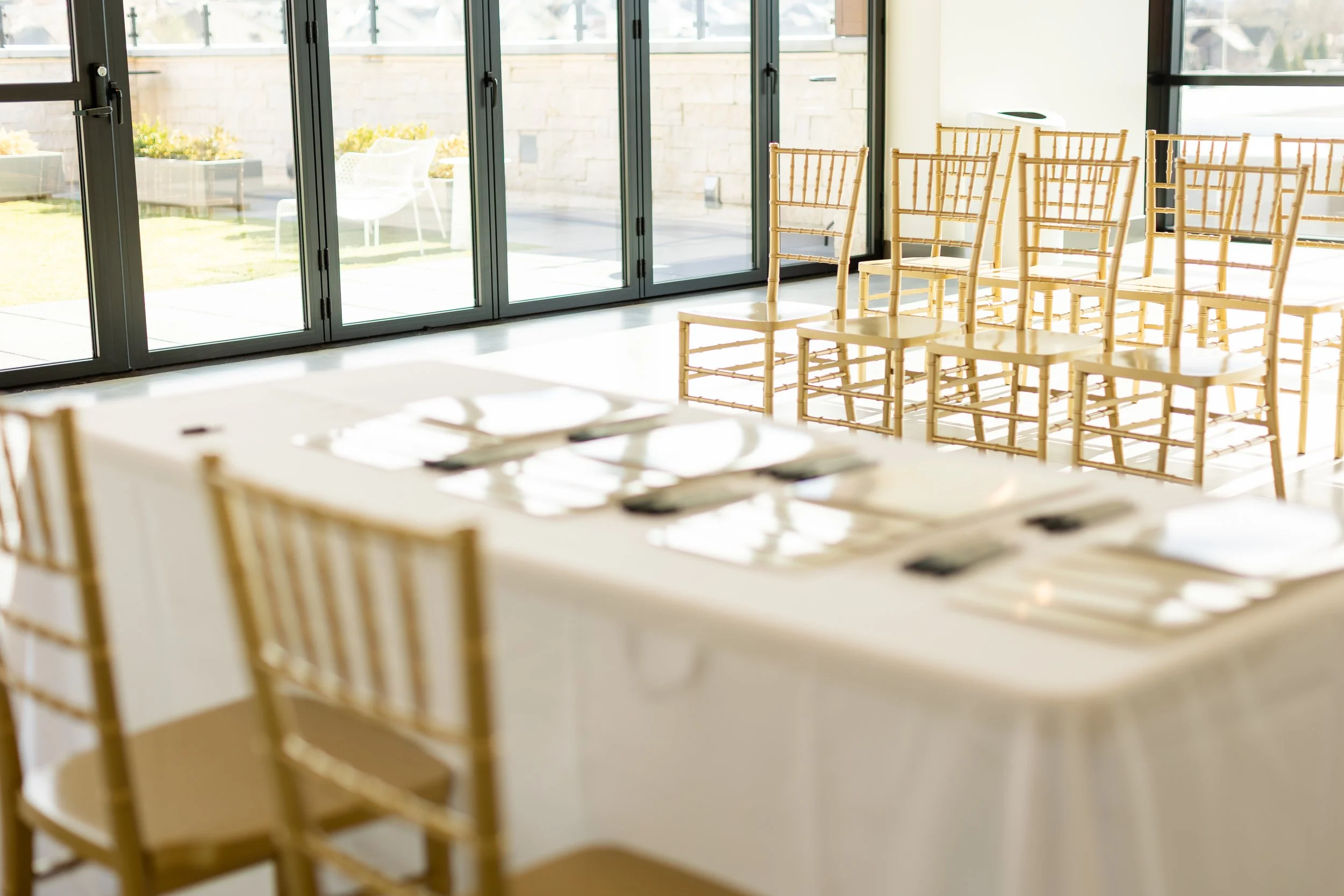 An indoor corporate event is setup with gold chairs facing a long rectangle table with gold chairs behind it.