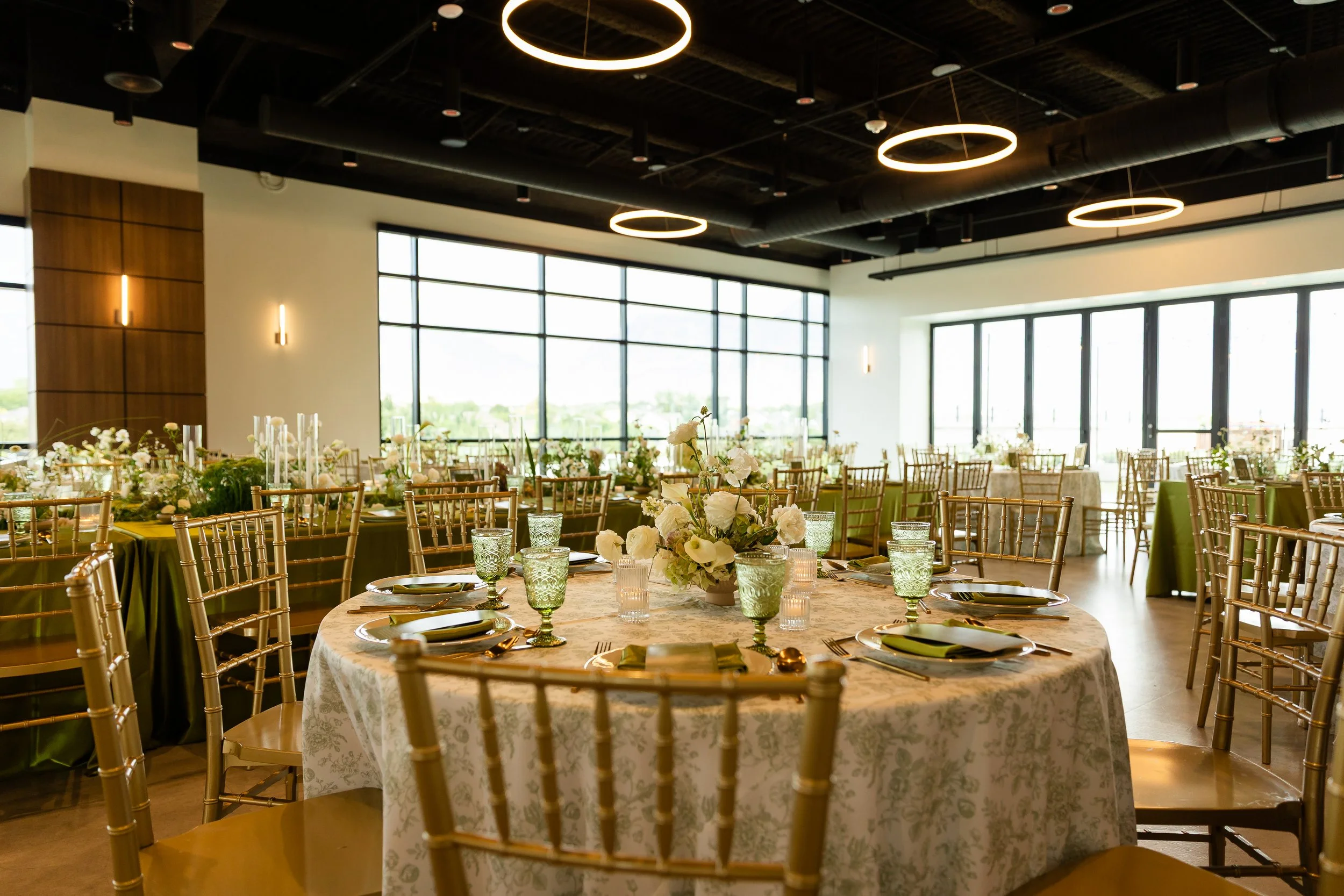 Round table set up for rehearsal dinner with a green and white floral linen, green goblets, and a floral centerpiece.