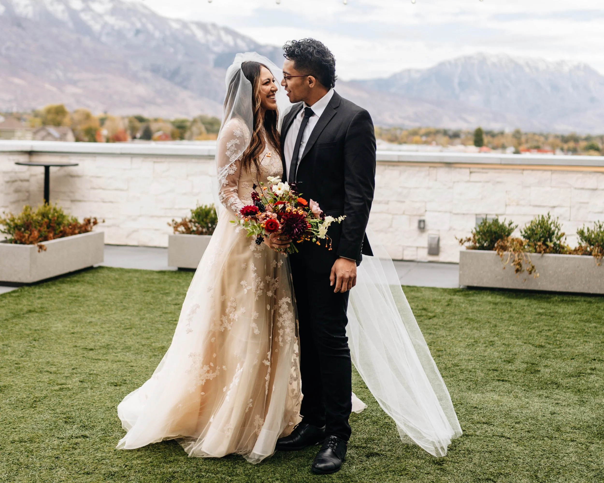 Bride and groom models stand next to each other in the courtyard looking at one another and smiling.