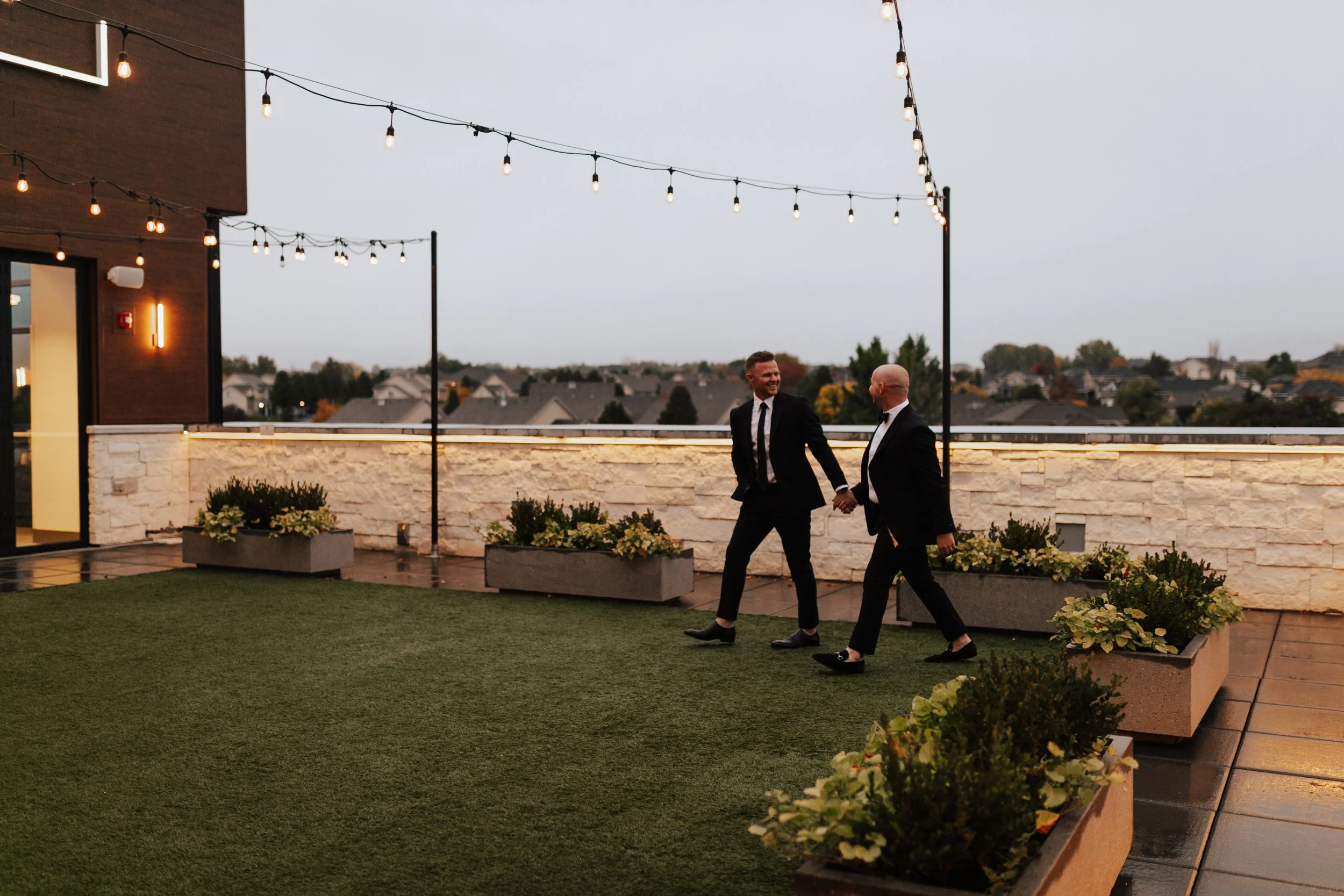 Two grooms wearing black suits walk across the outdoor courtyard holding hands.