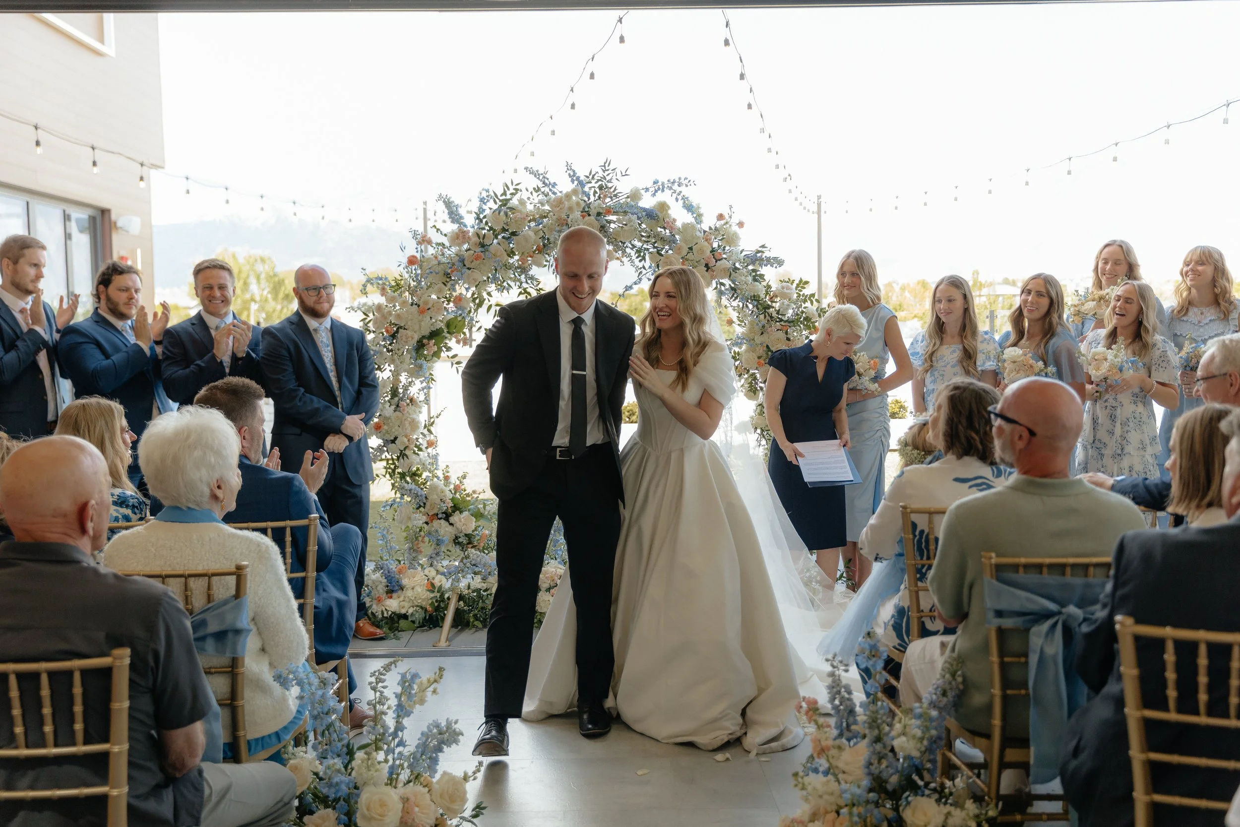 Bride and groom stand at the end of the wedding aisle smiling while the groomsmen, bridesmaid, and audience clap.