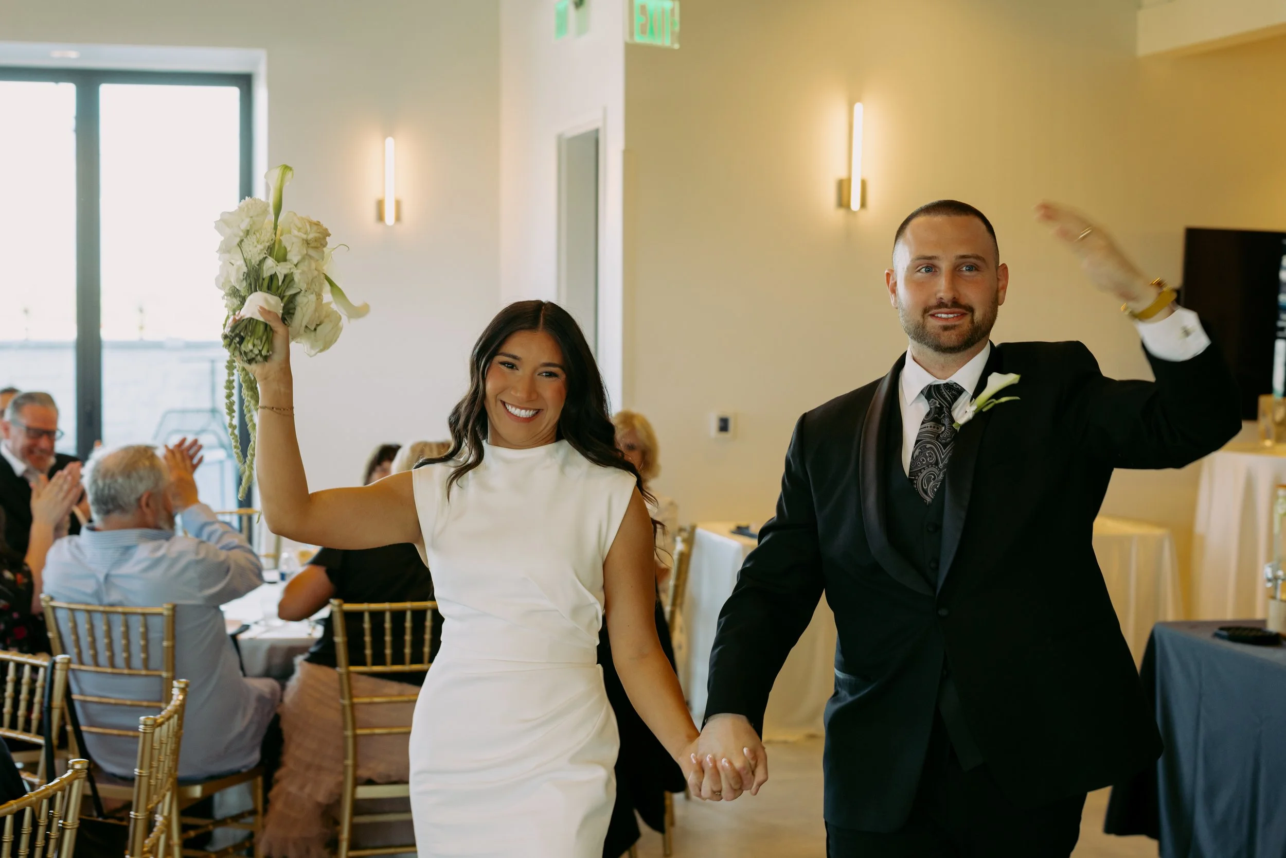 Bride and groom walk into the room holding hands and raising their other hands up to greet the crowd.