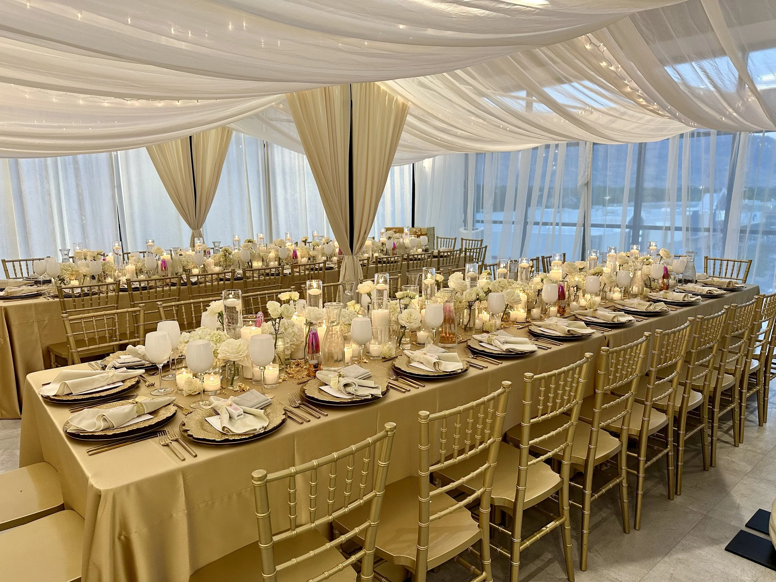 Long rectangle table setup with gold linens, white flowers, and white candles with sheer white drapes across the ceiling.