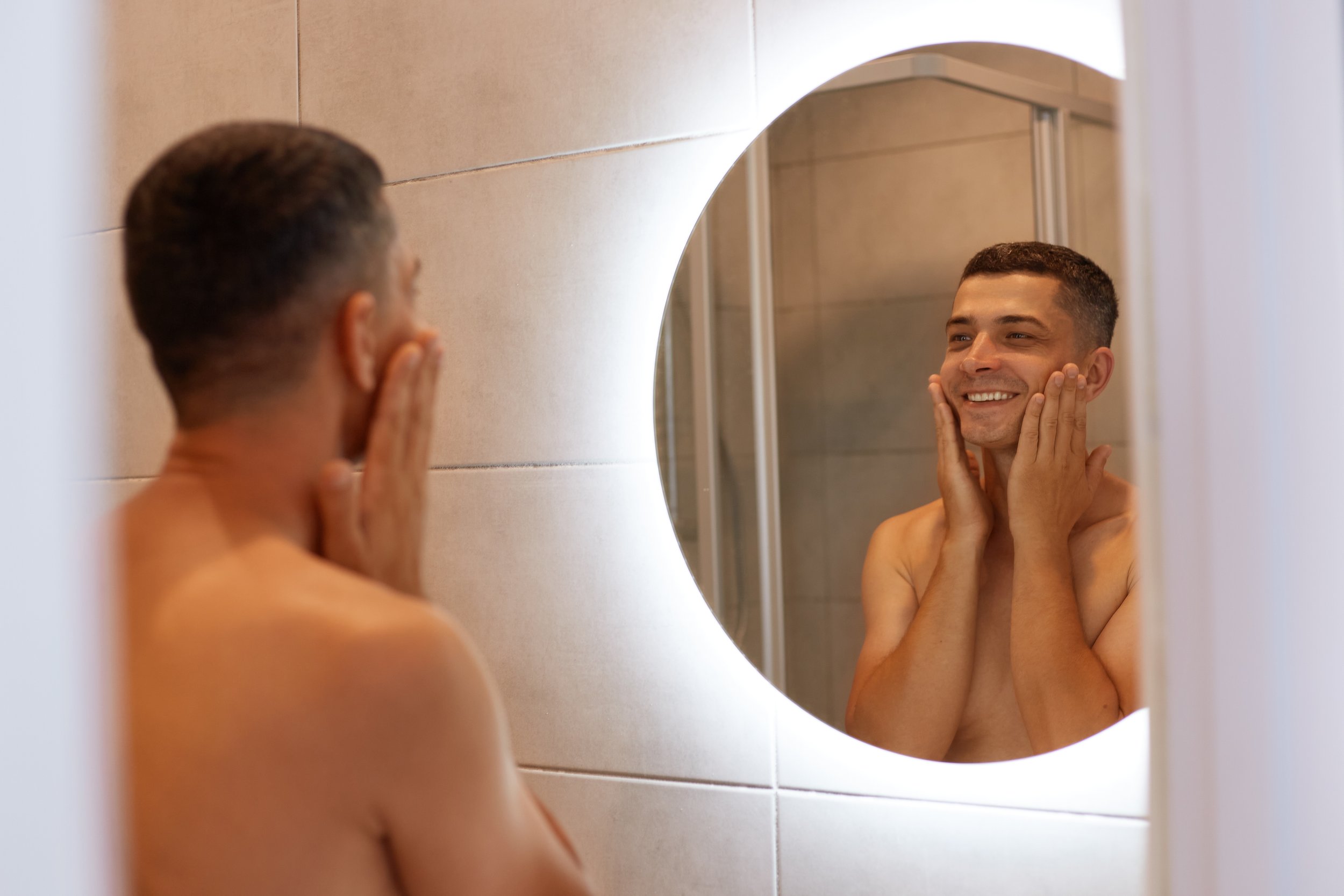 happy-positive-brunette-man-standing-bathroom-looking-his-reflection-mirror-touching-his-cheeks-applying-shaving-agent-face-smiling.jpg