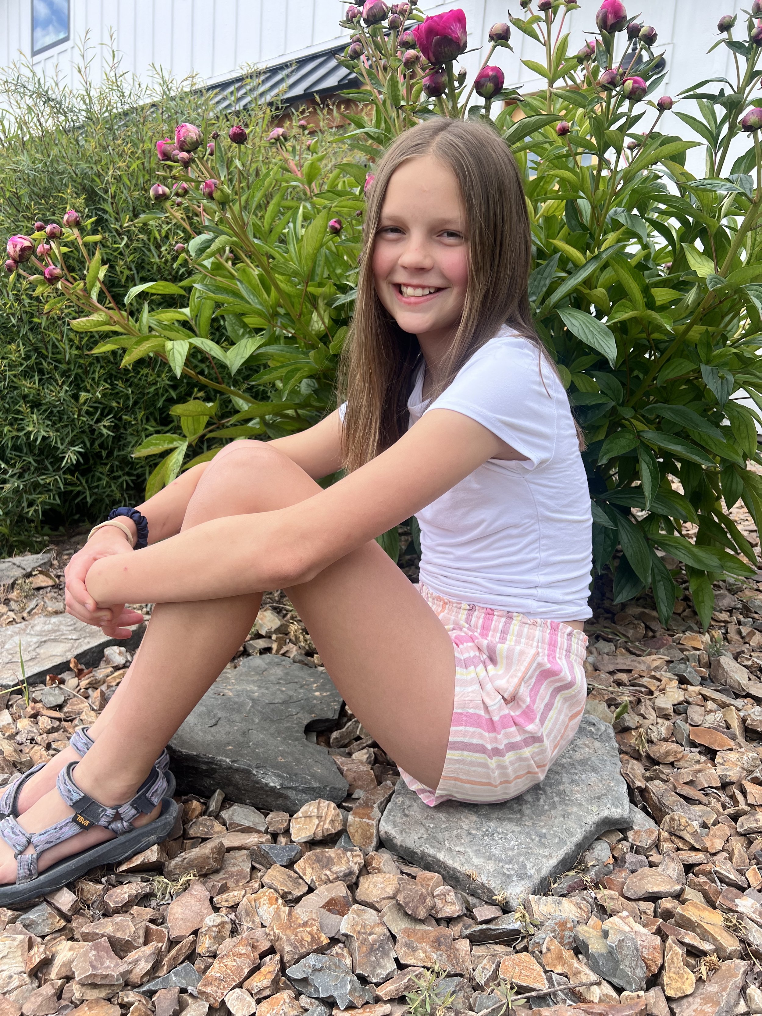 A young girl with long hair, wearing a white shirt and pink striped shorts, sitting on a rock amidst a garden of green plants and pink peonies, smiling at the camera.