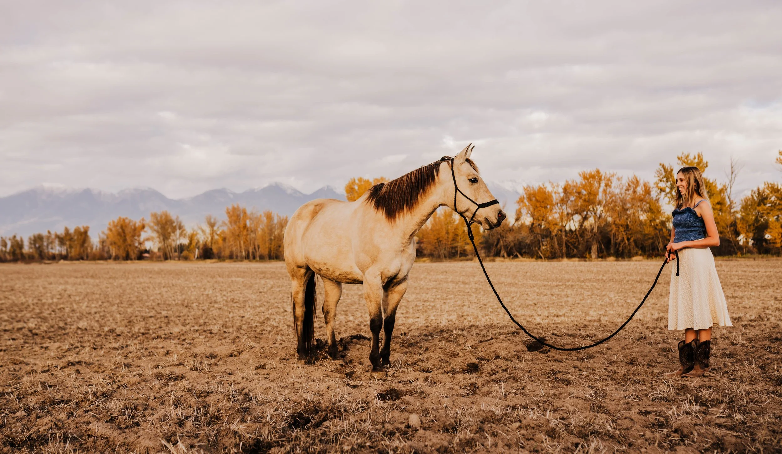 A woman with long hair wearing a blue top, cream skirt, and cowboy boots holds a black rope lead and smiles while standing on a dry, barren field with a light-colored horse. The horse wears a black halter and gazes at the woman. In the background, there are yellow-orange trees and snowy mountains under a cloudy sky.