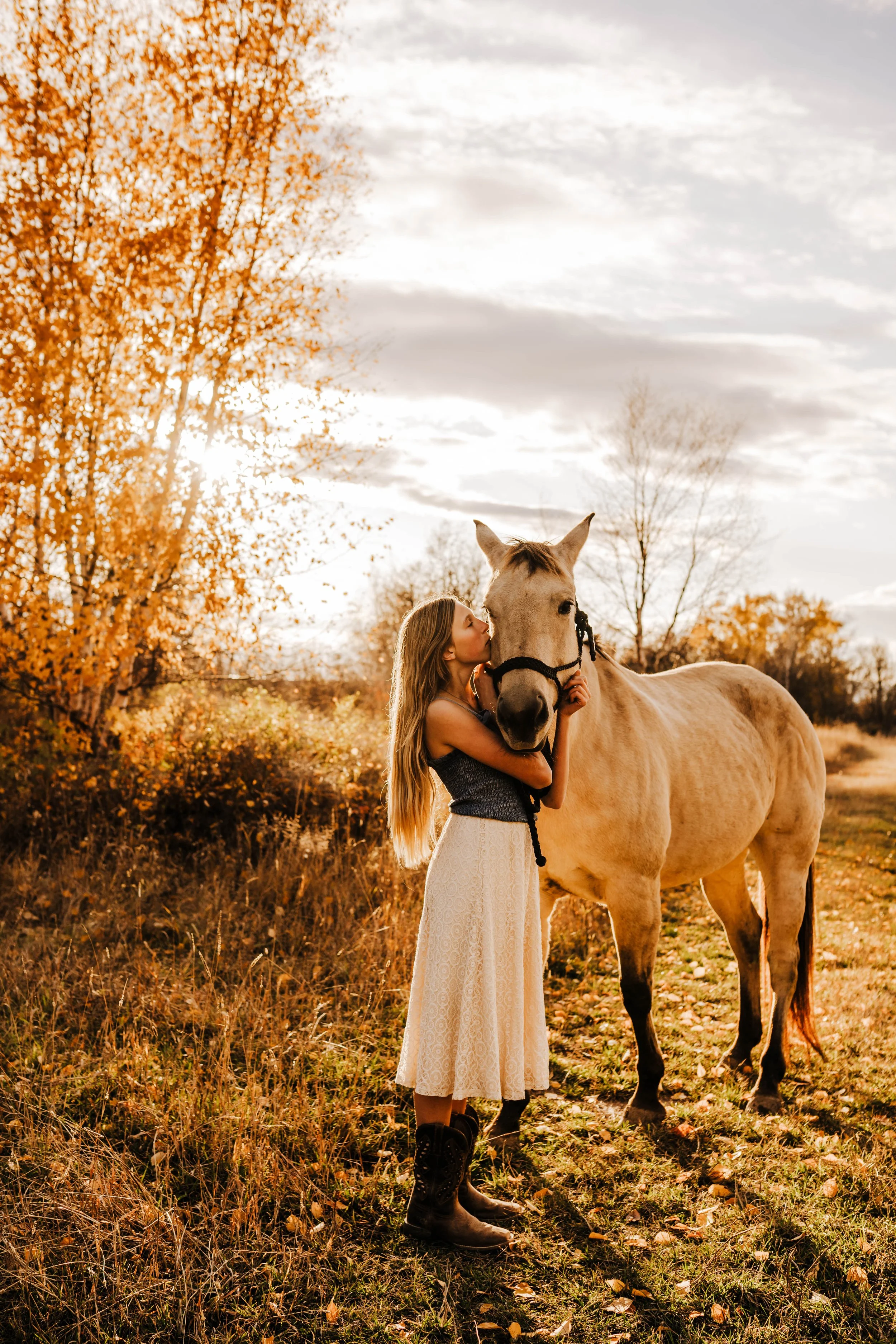 A woman with long blonde hair hugging a white horse in an autumnal landscape, with tall golden trees and a cloudy sky in the background.