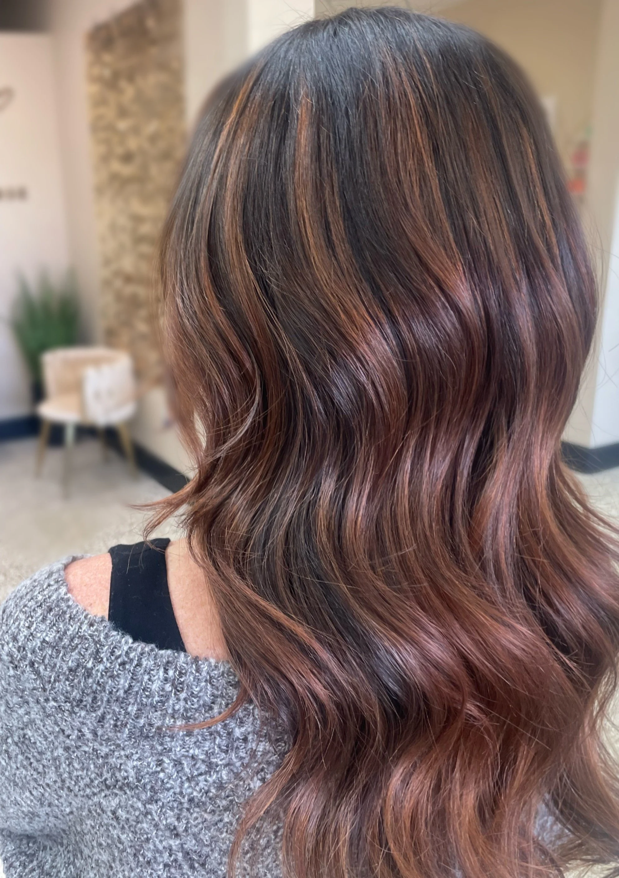 Woman with long wavy hair featuring brown and copper highlights, indoors with blurred background.