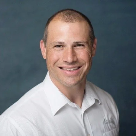 Man smiling in a white dress shirt against a dark blue background.