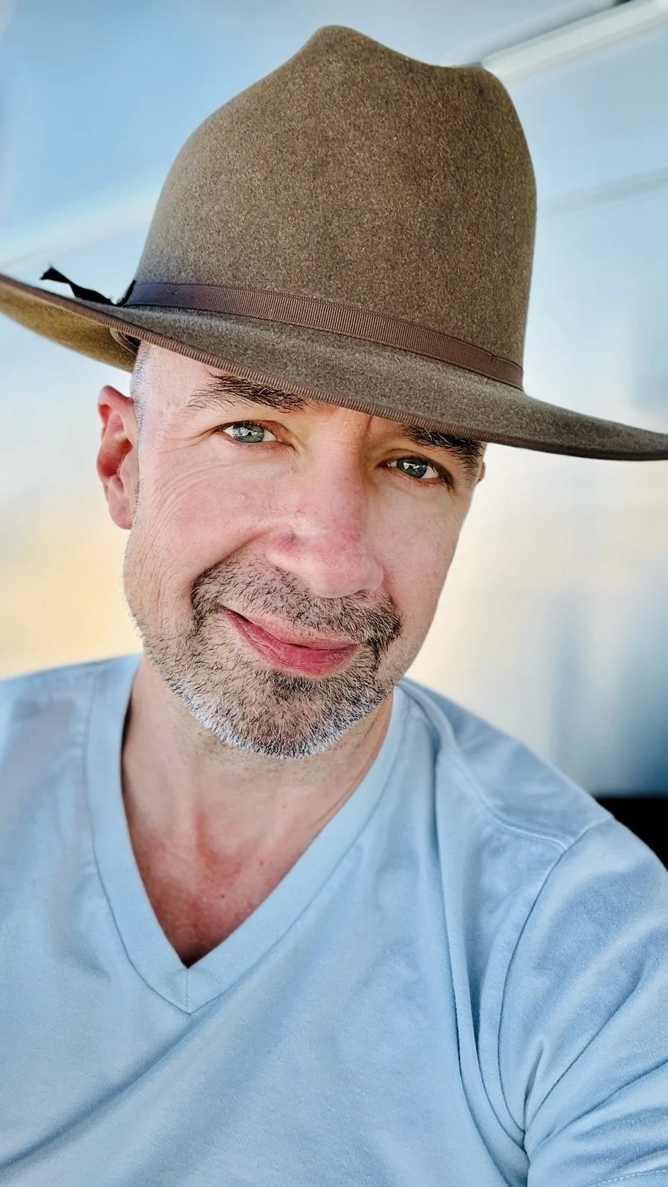 A man with a trimmed beard and blue eyes wearing a light blue V-neck shirt and a brown fedora hat, smiling outdoors.