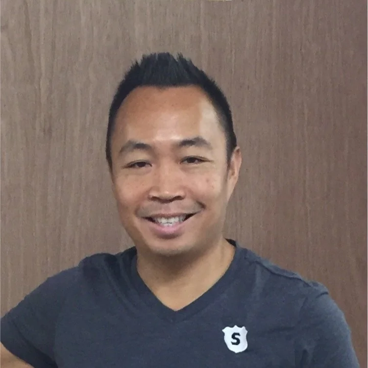Smiling person in a gray t-shirt with a small badge on it, standing against a wood-paneled background.