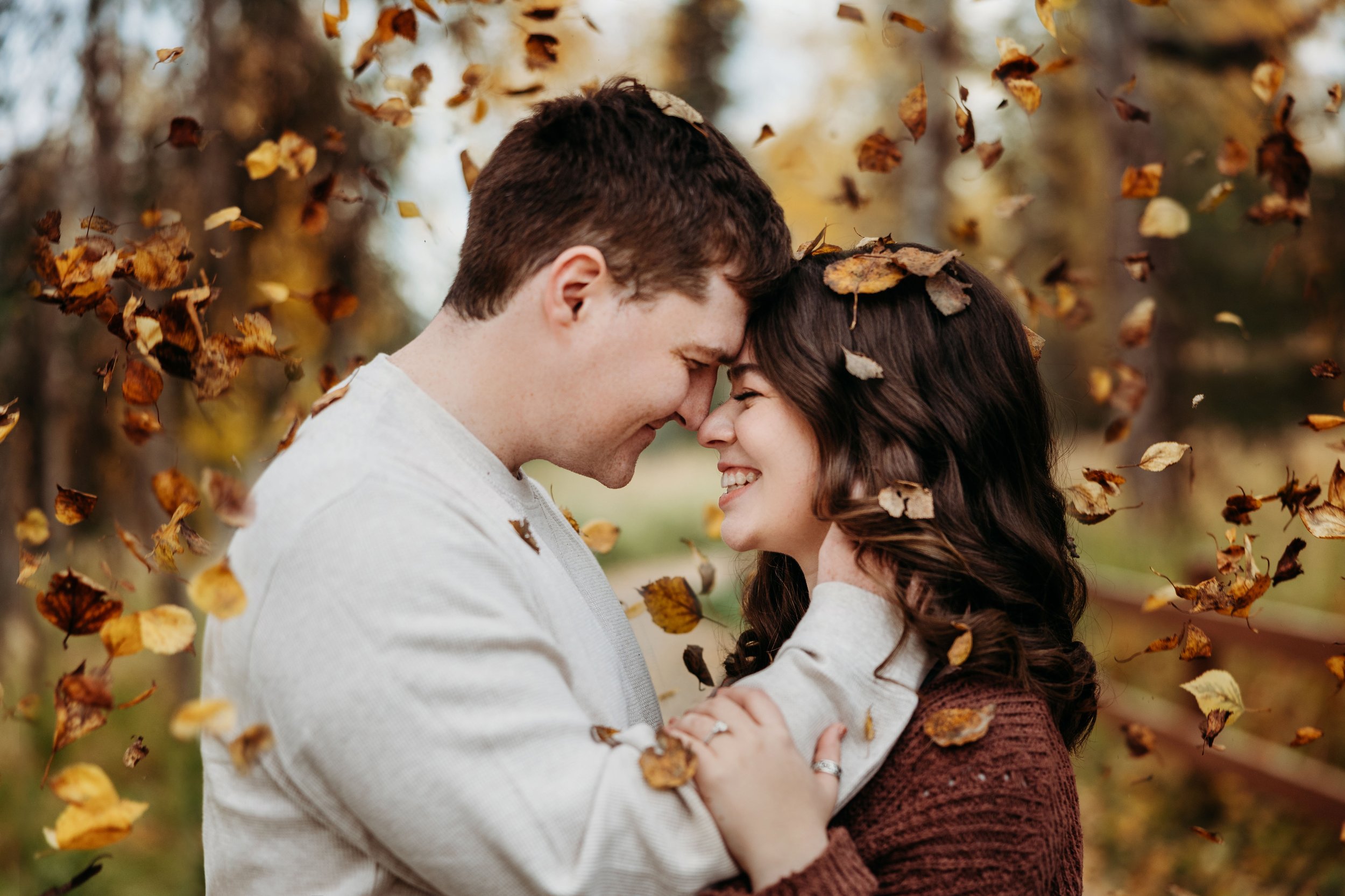 A couple smiling and embracing amid falling autumn leaves in a park setting.