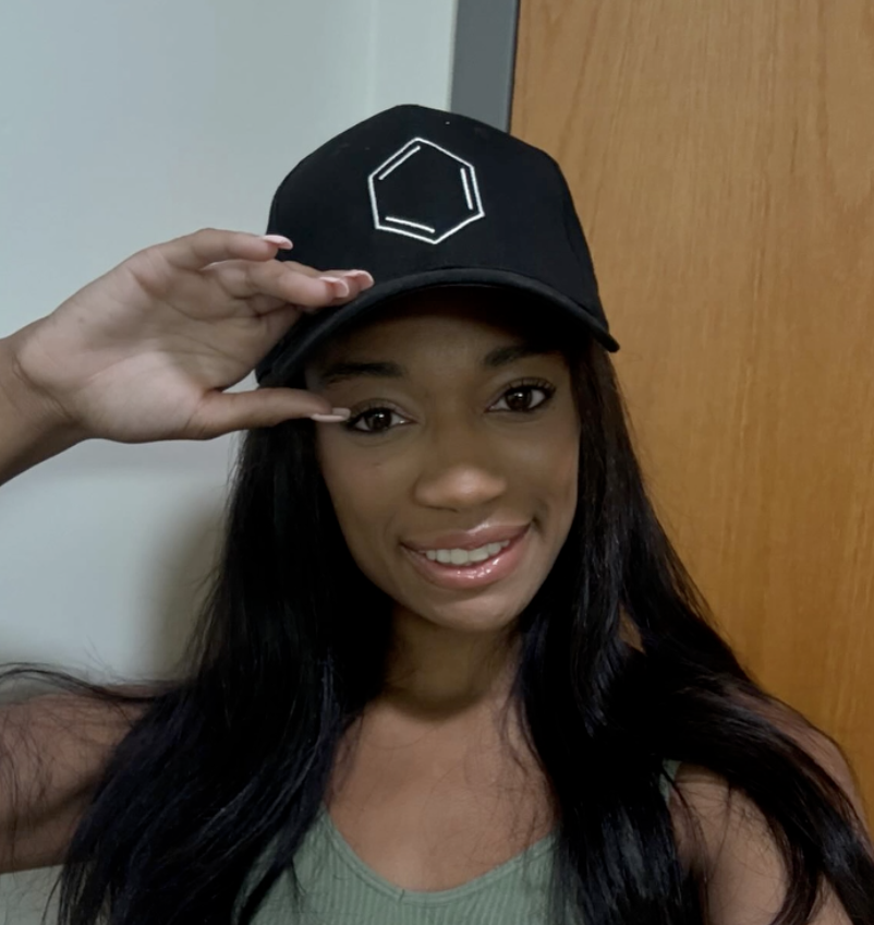 Woman smiling, wearing a black baseball cap with a white geometric design, and a green tank top, standing in front of a wooden door.