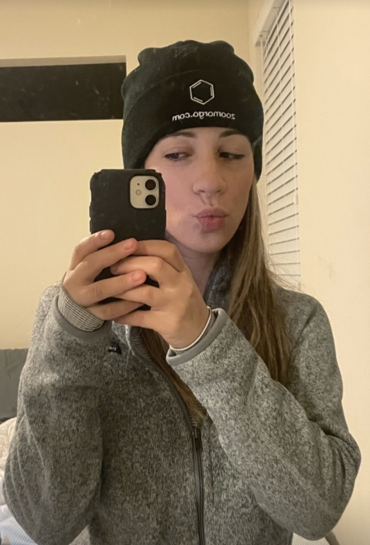 A young woman taking a mirror selfie, wearing a black beanie with a logo and website, a gray zip-up jacket, and making a slight duck face expression.