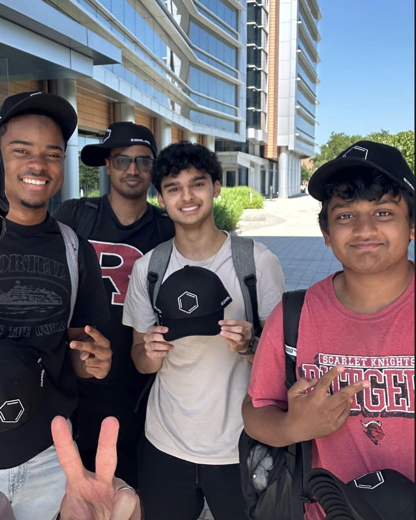 Four young men outdoors in front of a modern building, smiling and posing for a photo, some holding black hats with white designs, one making a peace sign, another pointing with two fingers.