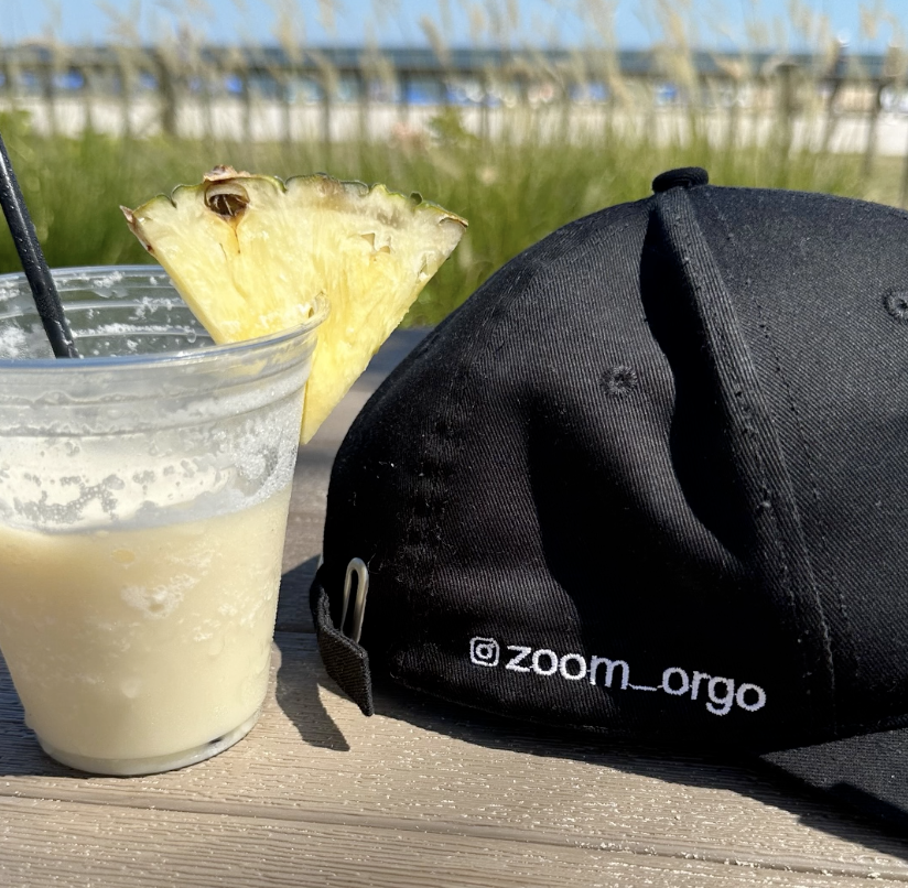 A glass of pineapple-flavored beverage with a pineapple wedge garnish, a black cap with the Instagram handle '@zoom_orgo', and a grassy background with a blue sky.