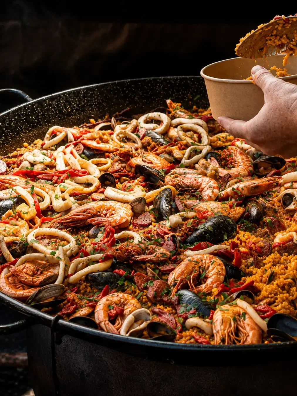 A hand scooping seafood paella loaded with prawns, mussels and calamari into a takeaway bowl at Eat Drink Nights in Kirrawee.