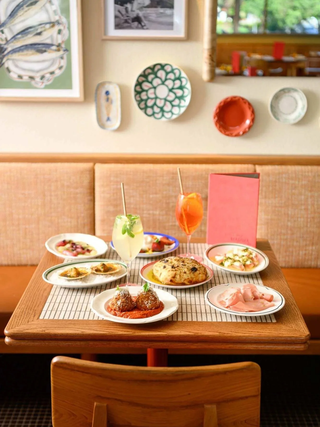 A colourful spread of Mediterranean-inspired dishes and cocktails at Call Me Mumma in Parc, Cronulla, featuring wooden tables, peach-toned booth seating, decorative ceramics and vibrant wall art.