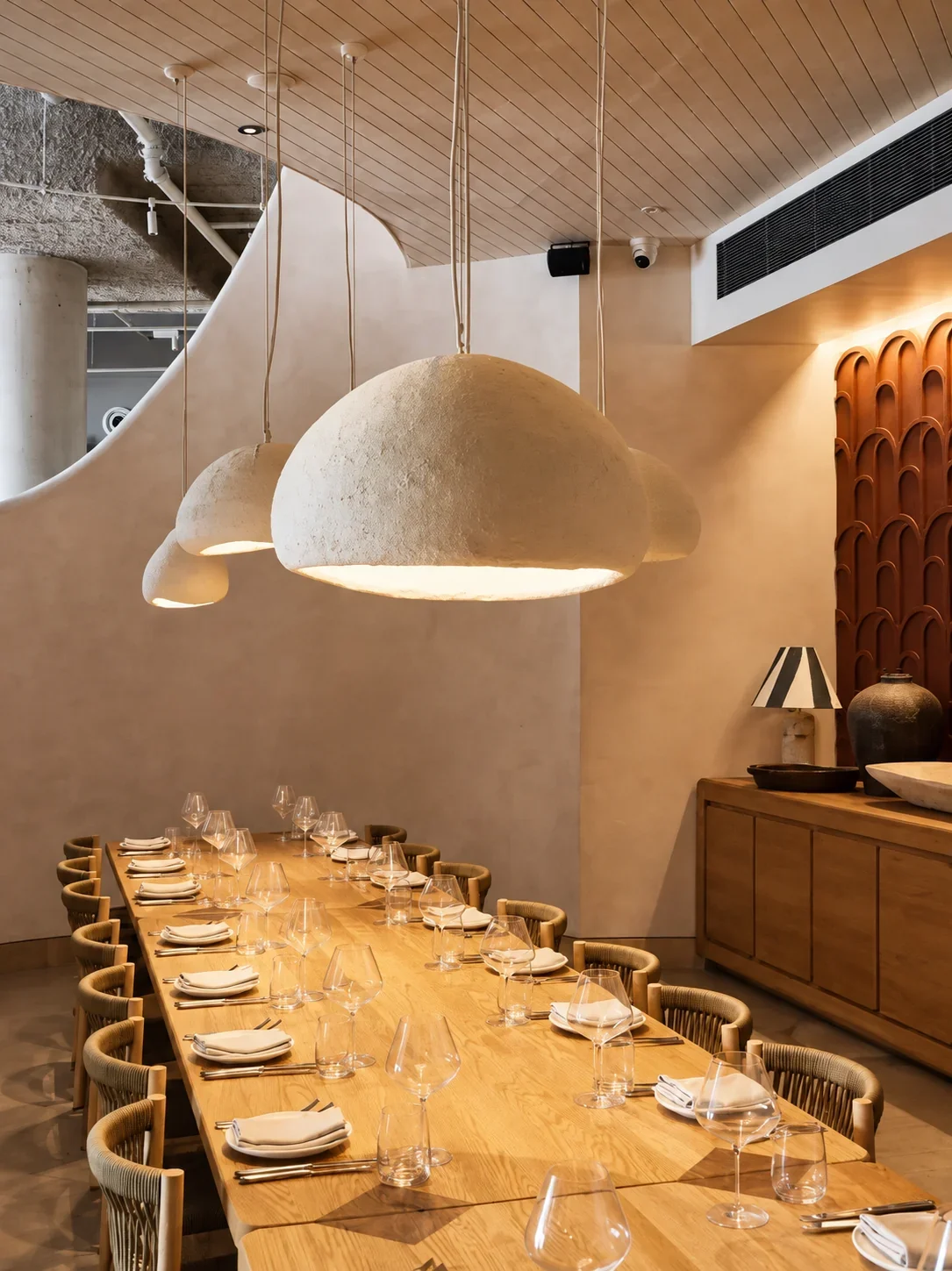 Elegant dining space at Hurricanes Cronulla, featuring a long timber table set with glassware and white napkins, sculptural pendant lighting, curved walls and warm neutral interiors.