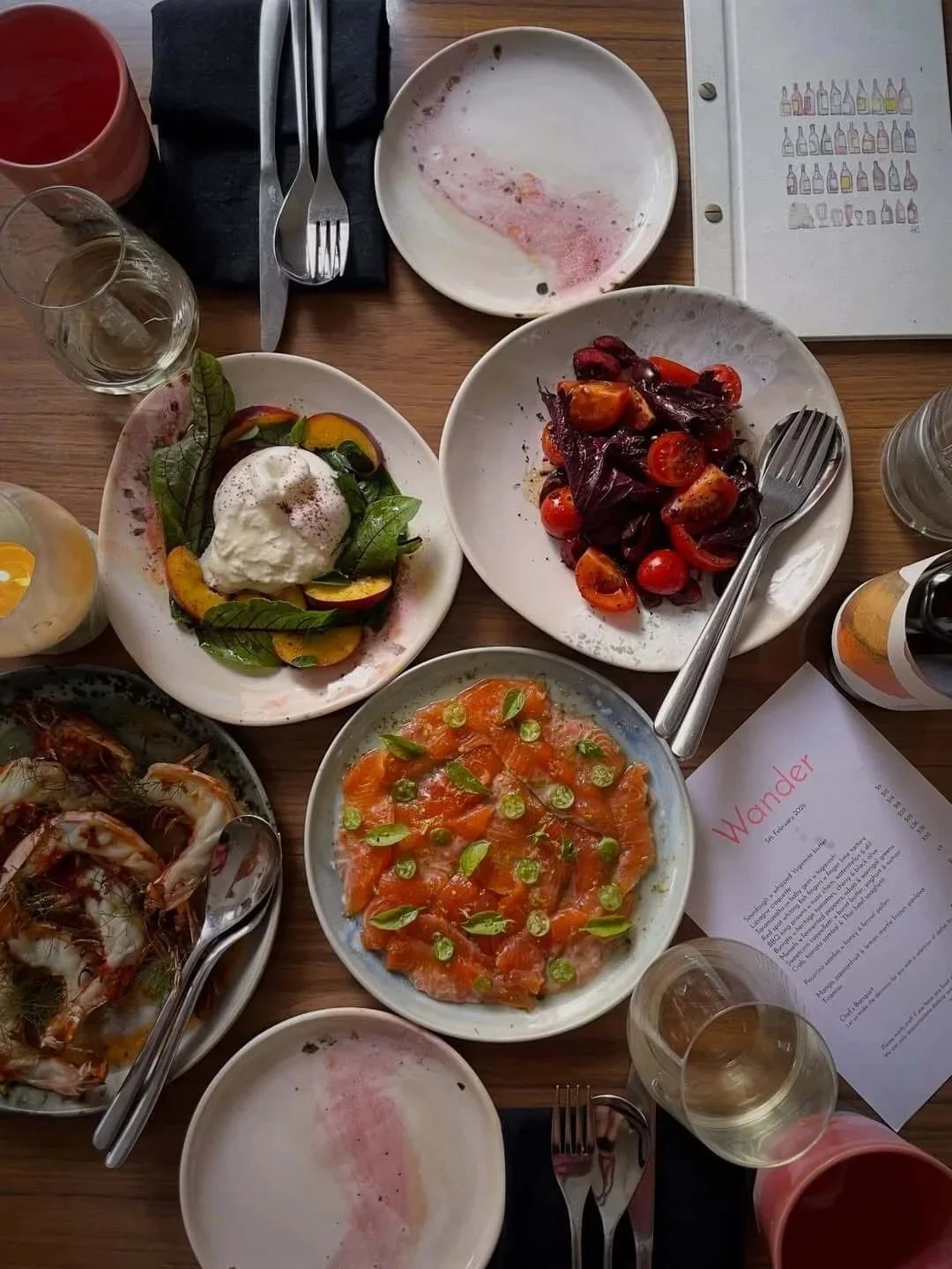 an overhead shot of a shared meal on a timber table with speckled ceramic plates featuring burrata with peach and basil, salmon carpaccio with sliced chilli, a tomato and radicchio salad, grilled prawns, water glasses and a Wander restaurant menu.