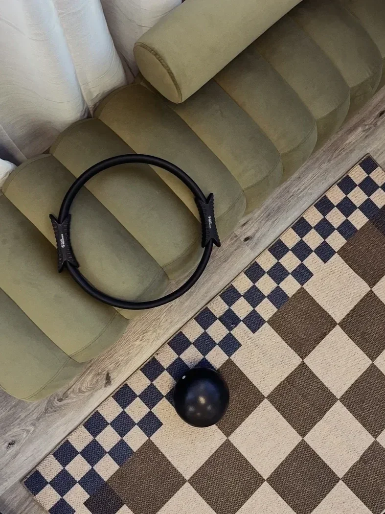 green upholstered bench above a checkered rug with a pilates strength ring on top of it