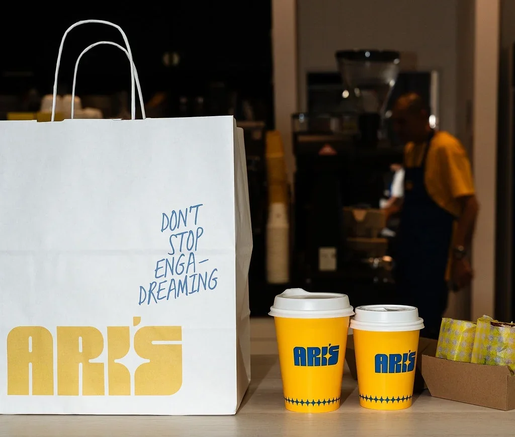 A white ARI'S branded takeaway paper bag with blue handwritten text beside two yellow ARI'S coffee cups with white lids, set on a wooden café counter with a barista working in the background.