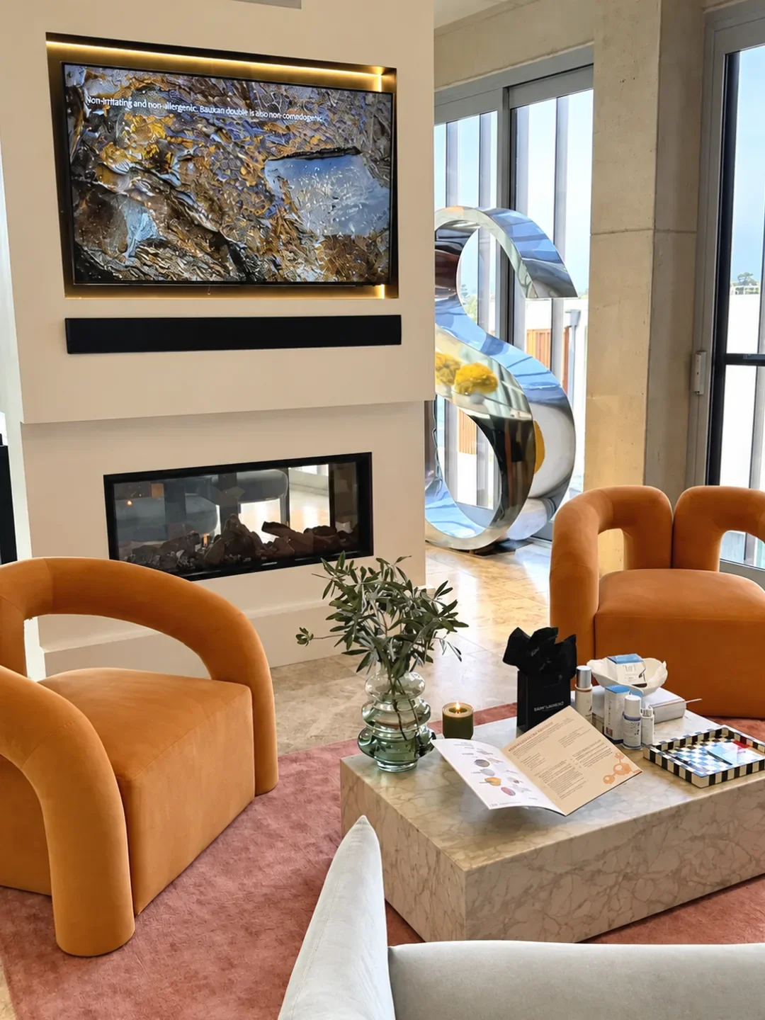 A lounge at Skin Street Clinic in Kangaroo Point with orange velvet armchairs, a fireplace and skincare products on a marble table.