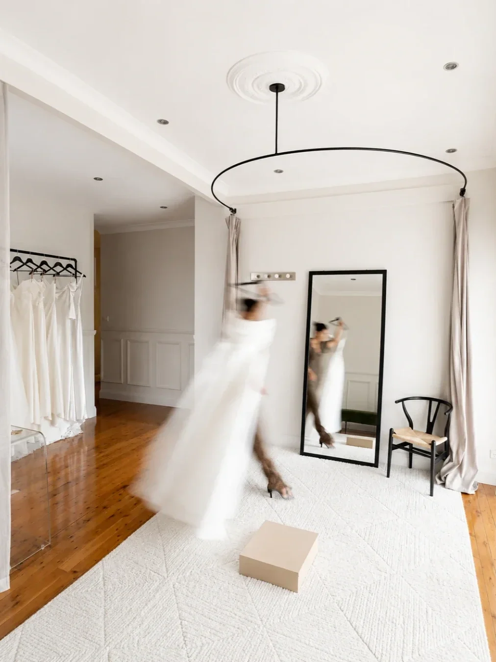 Bright bridal boutique interior with white wedding dresses on display and a blurred figure in a flowing gown moving in front of a full-length mirror.