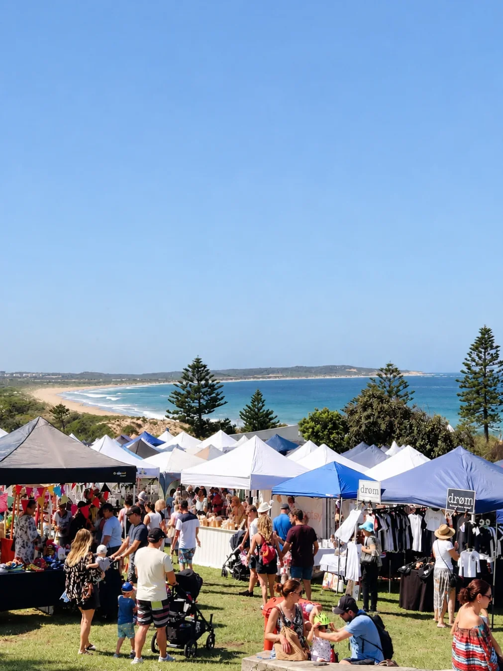 Crowds browsing white and blue market stalls on a grassy headland in Cronulla, with the beach and ocean behind.