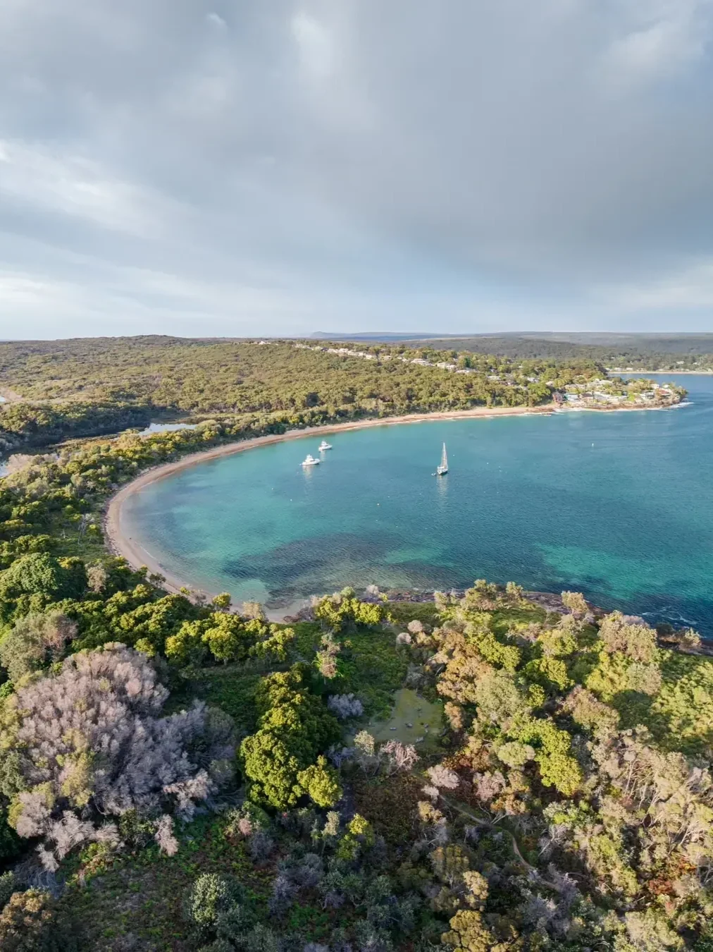 Aerial view of Jibbon Beach in Bundeena, Sydney, featuring a secluded crescent-shaped sandy beach, clear turquoise waters with sailboats and lush native bushland surrounding the peaceful coastal bay.