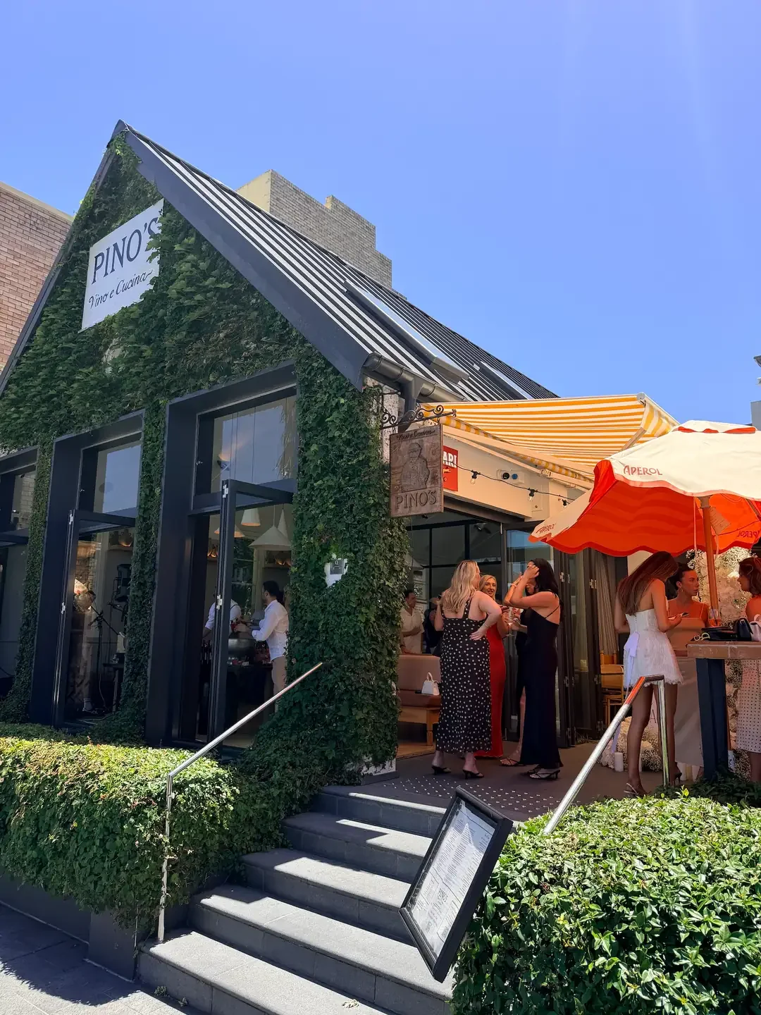 Exterior of Pino's Italian restaurant, featuring an ivy-covered façade, striped awnings and outdoor dining area with guests enjoying drinks beneath orange umbrellas on a sunny day.