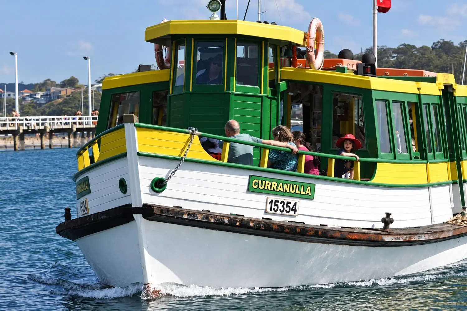 The green and yellow timber ferry Curranulla carries passengers past a wharf with Cronulla Ferries.