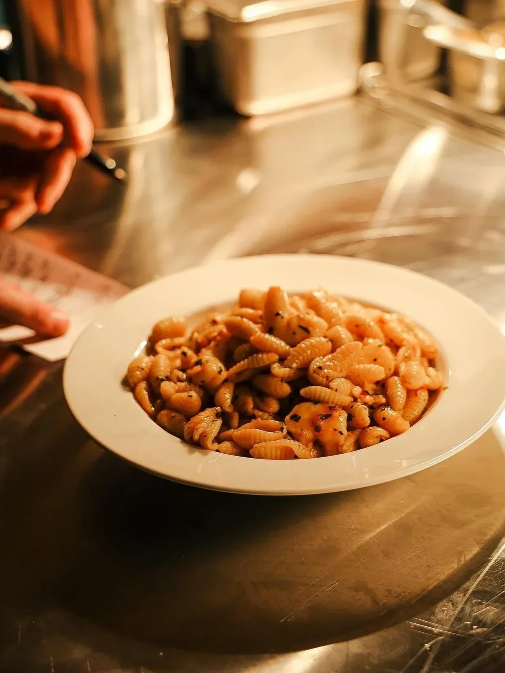 Close-up of a freshly plated pasta dish on a stainless steel kitchen bench, with a chef finishing the dish in the background.