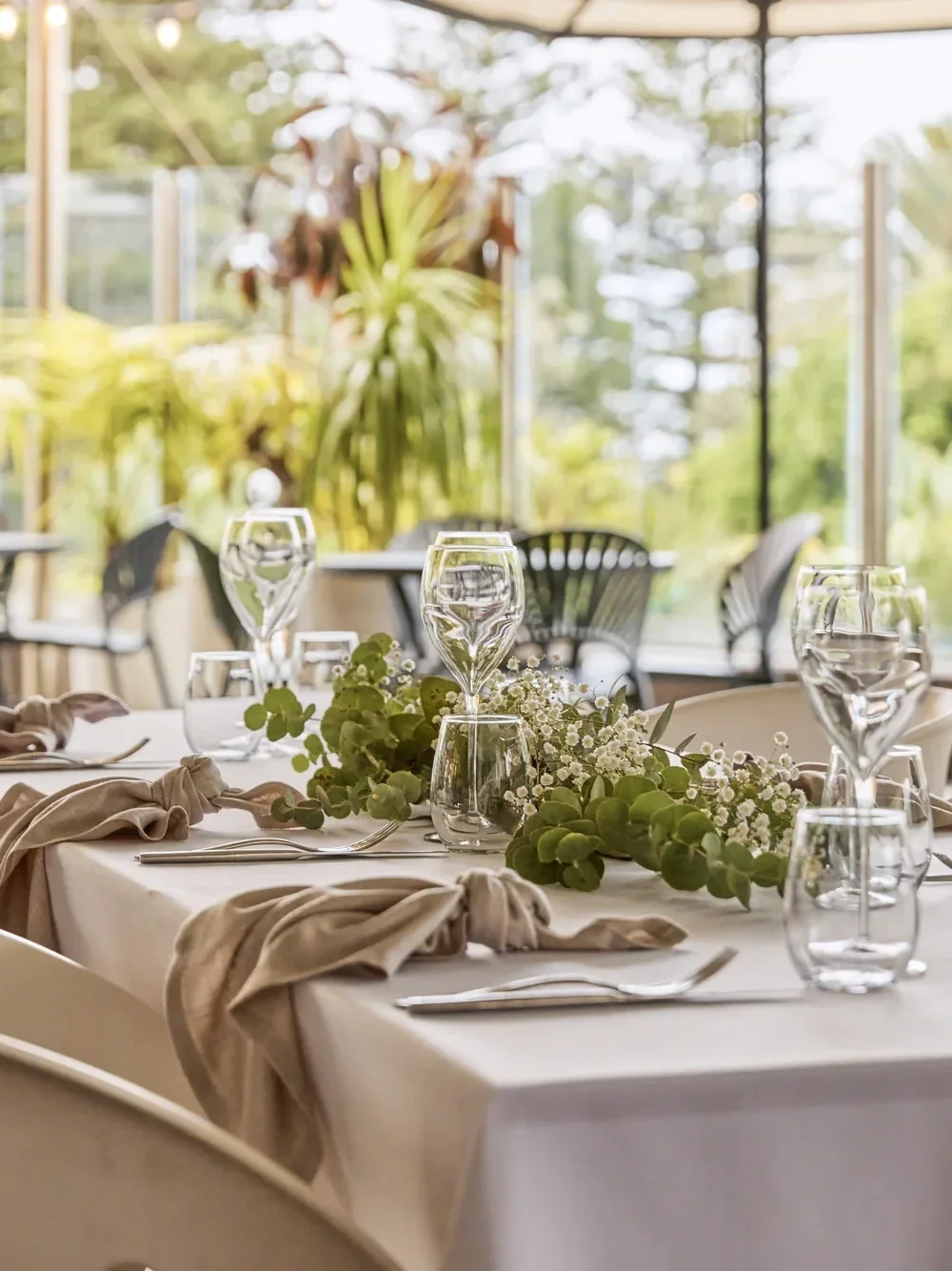 Elegant table setting at Rydges Cronulla's The Alley, featuring white tablecloths, neutral linen napkins, floral centrepieces and glassware, with a sunlit outdoor terrace and lush greenery in the background.