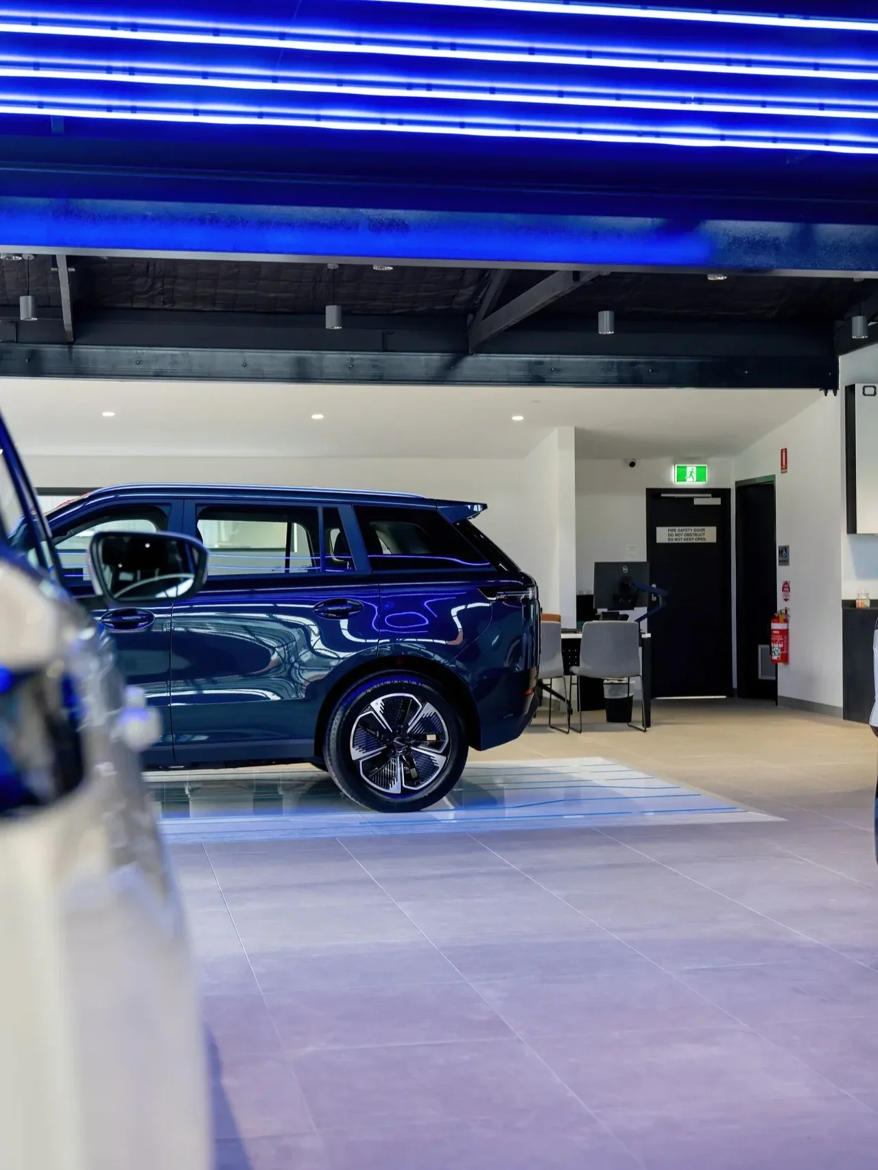 Blue SUV displayed inside a modern car dealership showroom beneath bright blue ceiling lights, with another vehicle blurred in the foreground.