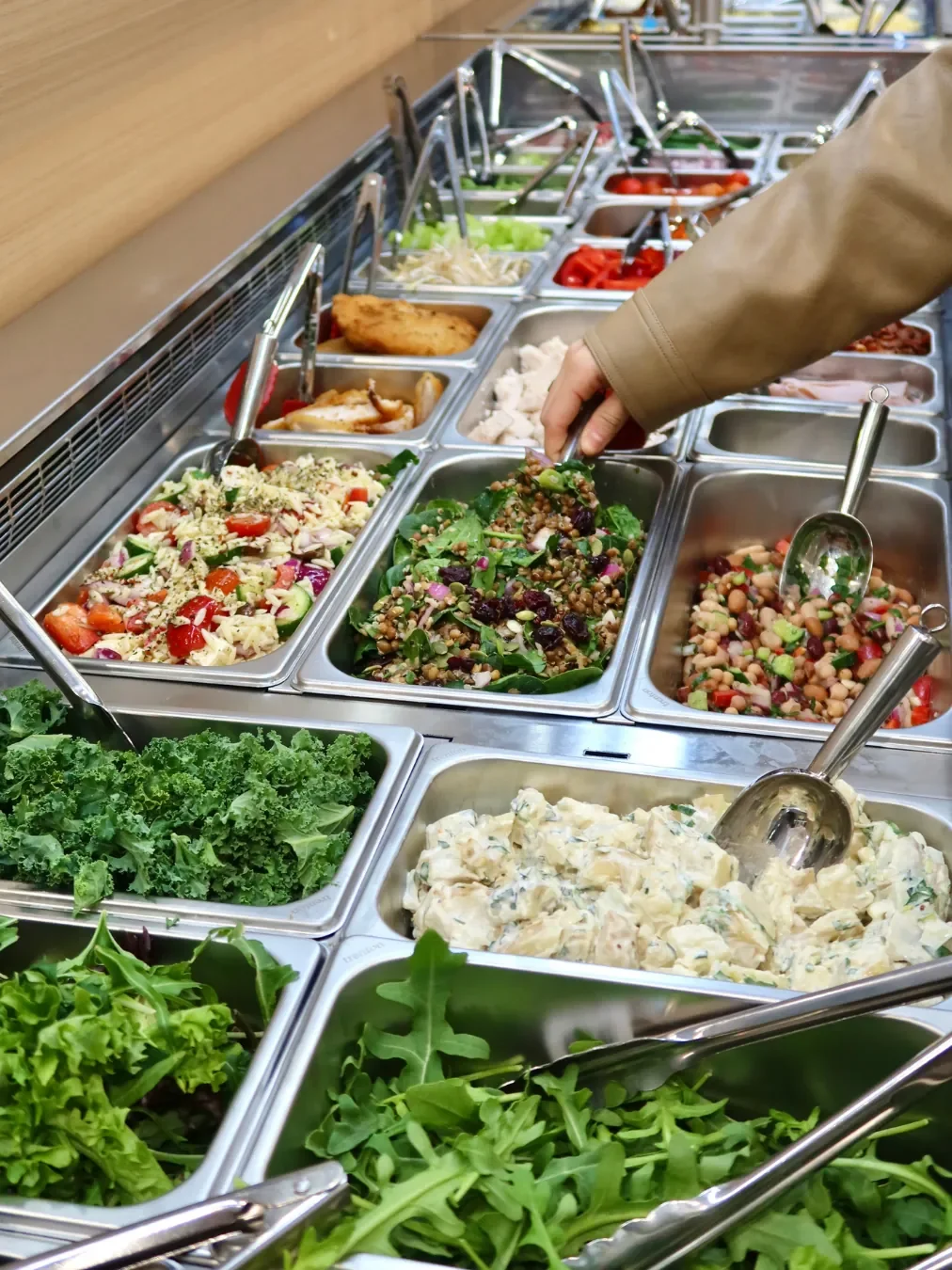 A hand serving from a self-serve salad bar filled with stainless steel trays of grain salad with cranberries, three-bean salad, potato salad, kale, rocket and mixed leaves.