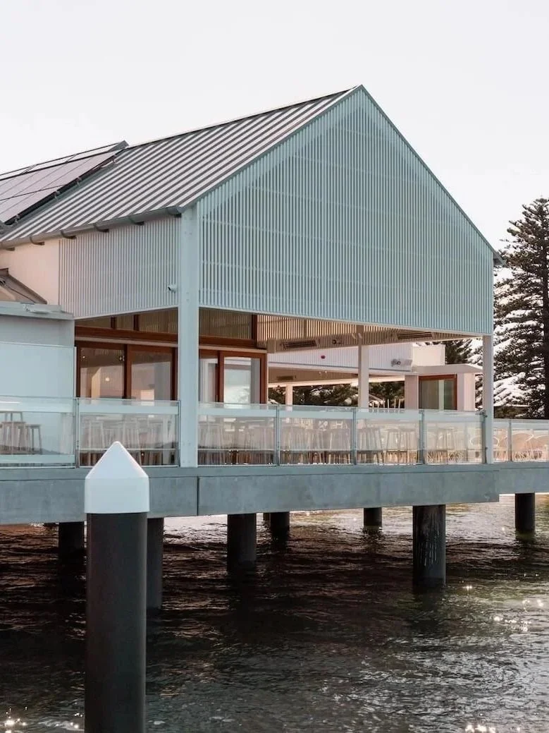 Exterior of St George Sailing Club, featuring a modern waterfront building with a pitched metal roof, white cladding and a spacious outdoor terrace overlooking the water.