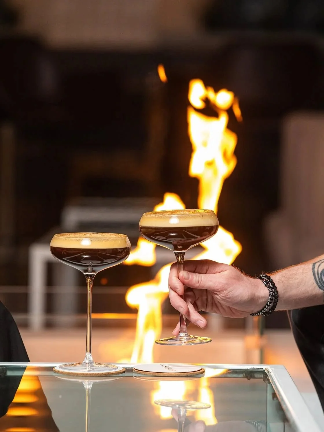 Two espresso martinis in coupe glasses, with one being held up in front of a glowing fireplace.