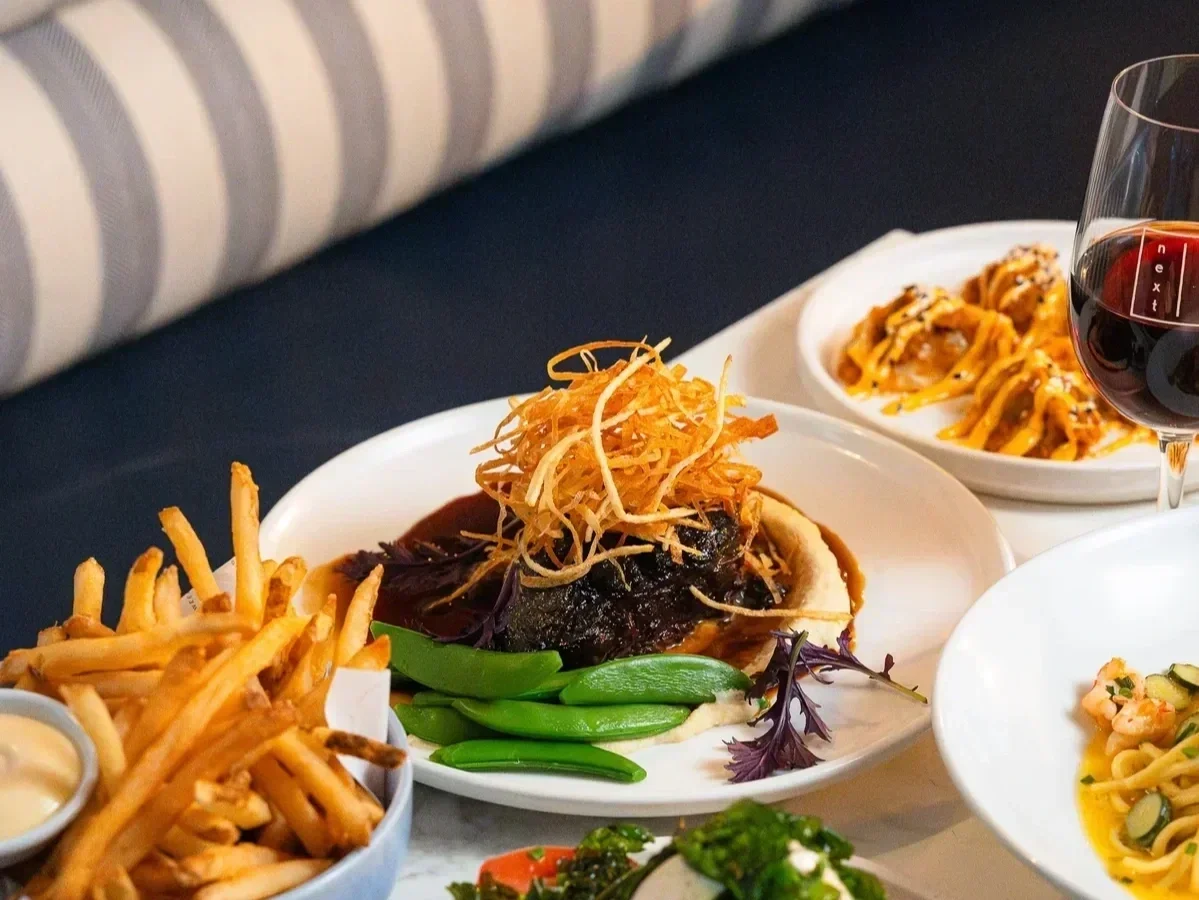 A restaurant table with a striped navy and white banquette, set with a bowl of golden fries, a braised short rib topped with crispy fried onions and served with sugar snap peas, a glass of red wine, and plates of pasta and dumplings in the background