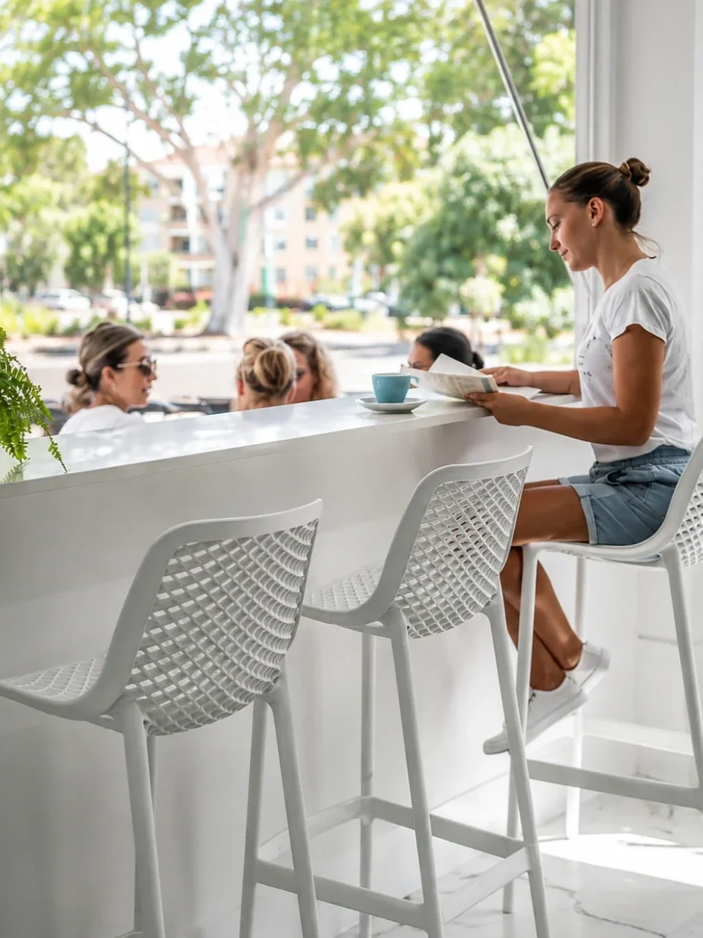 Woman sitting at a bright café window counter, reading a menu beside a cup of coffee, with other diners seated outside.