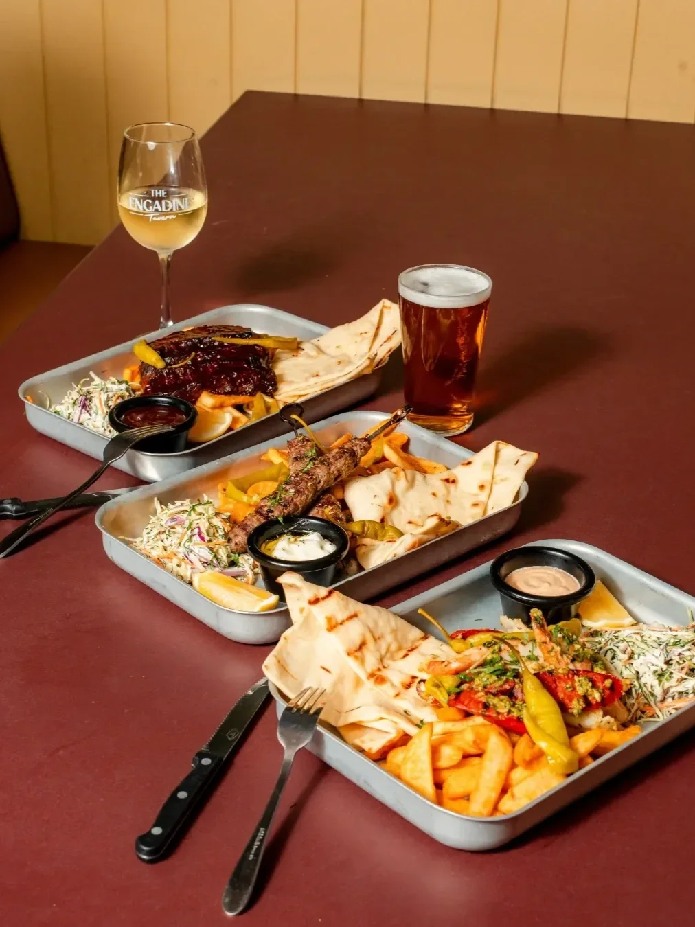Three pub meals with grilled meats, chips, flatbread, coleslaw and dipping sauces, served alongside beer and white wine.