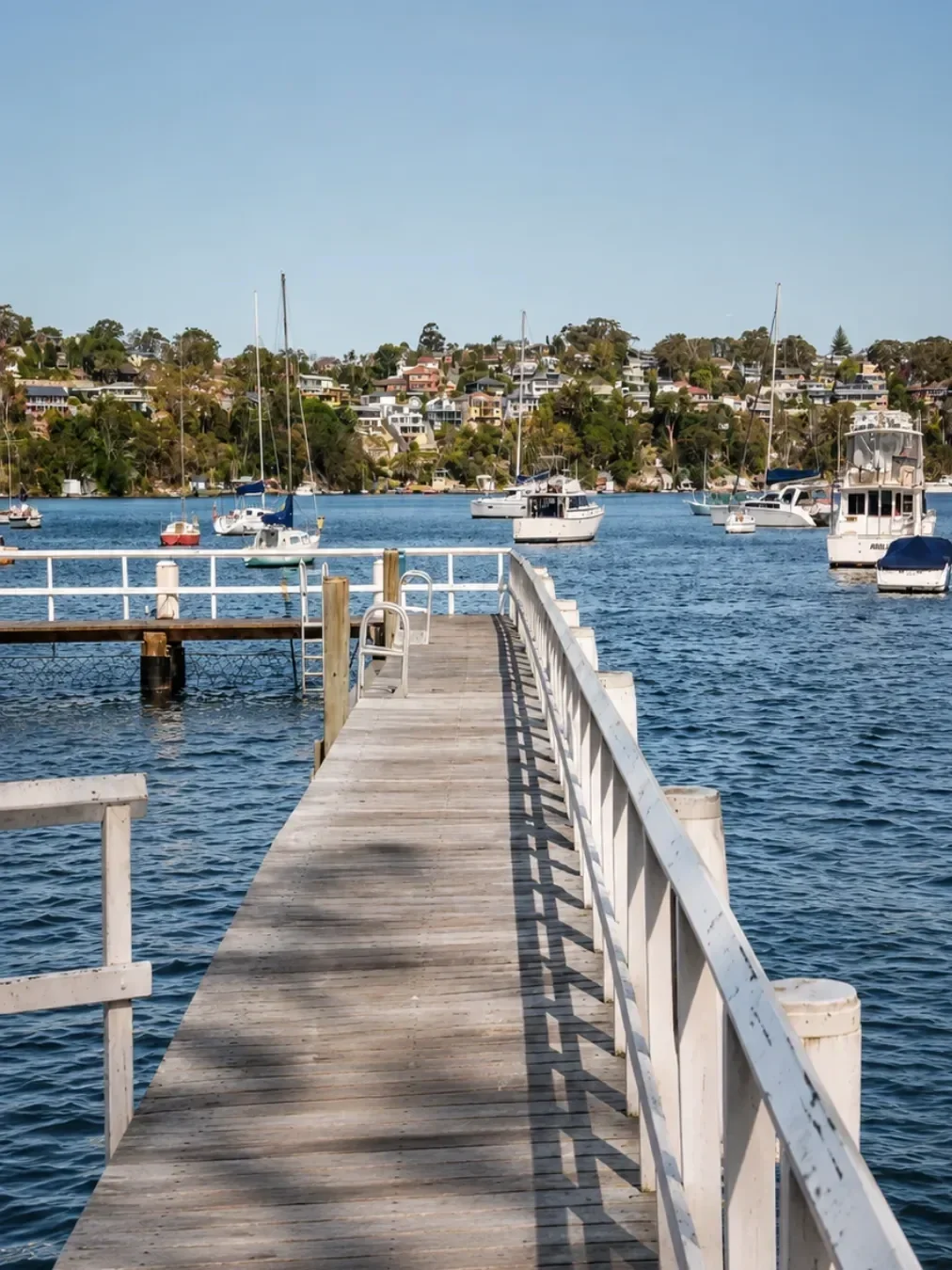 Wooden jetty extending into Gymea Bay, with boats moored on the water and waterfront homes lining the opposite shore.