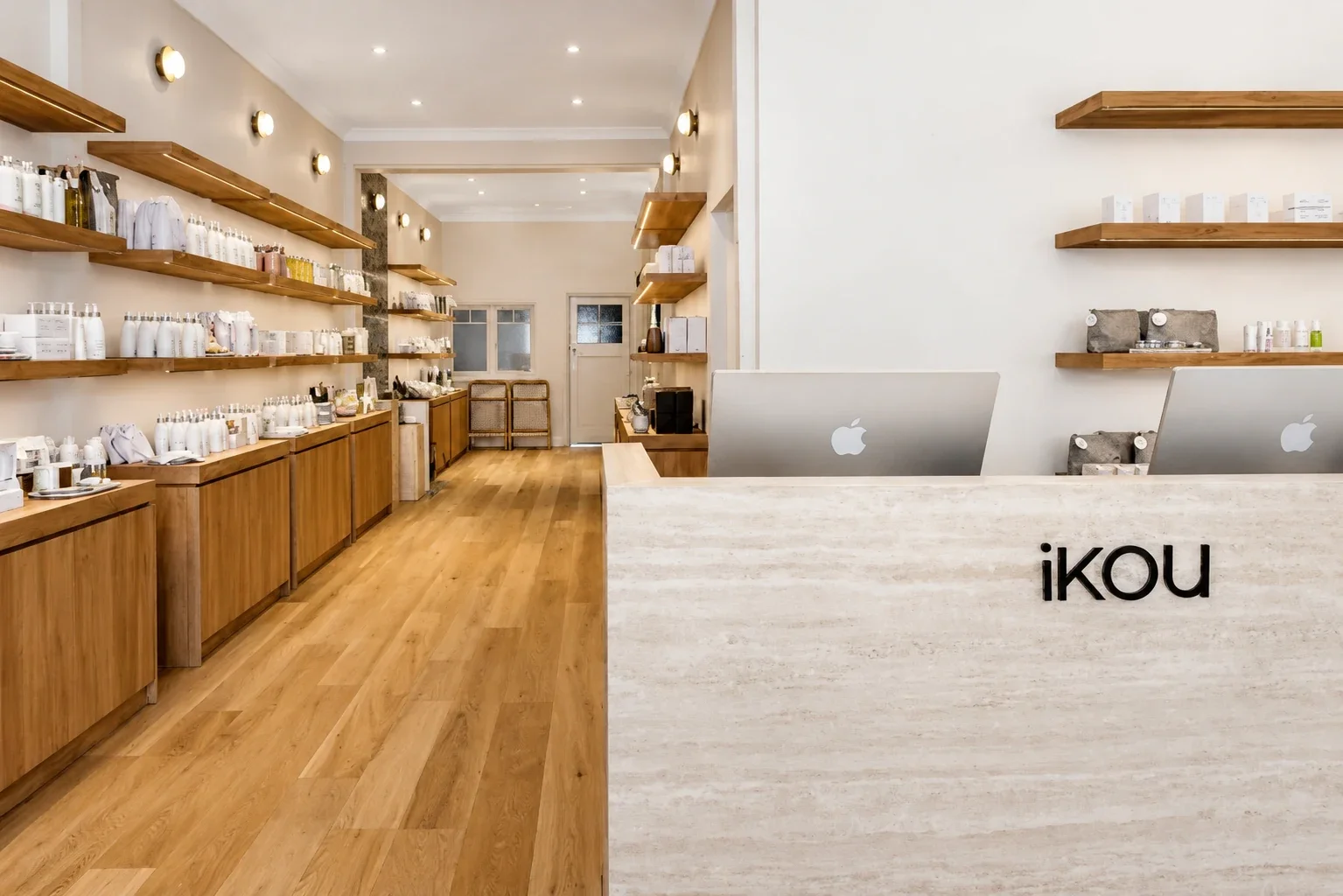 Interior of iKOU store in Berry, NSW, with timber shelving of skincare and candle products, oak flooring, and a travertine reception counter with iKOU signage and iMac displays