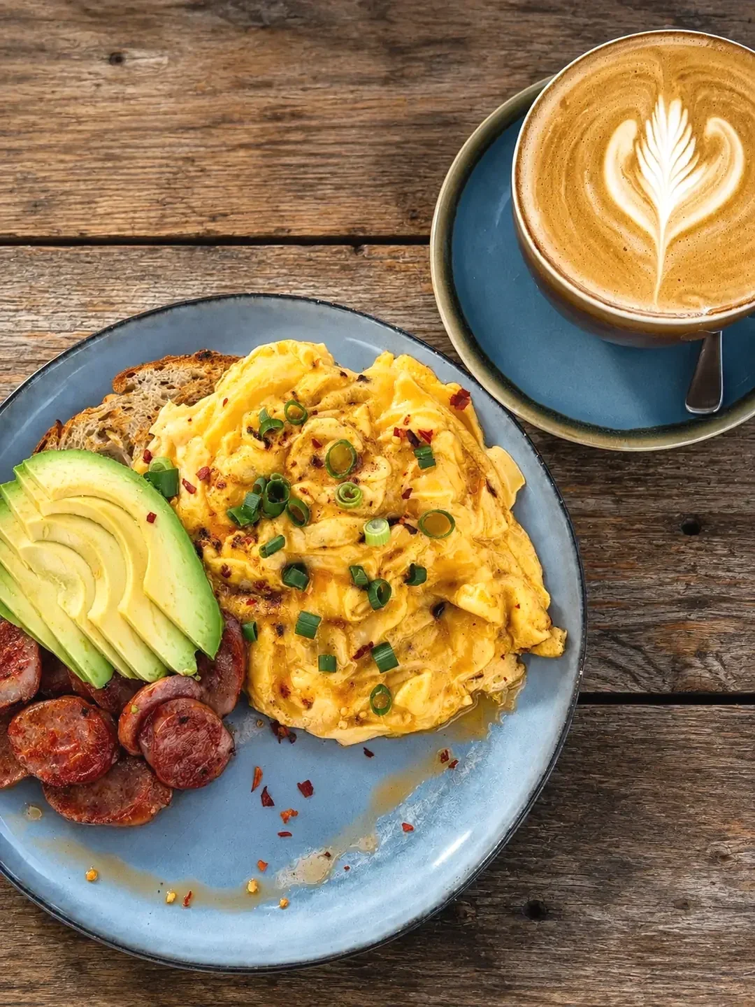 Scrambled eggs with chives, sliced avocado, chorizo and sourdough served beside a latte on a wooden table.