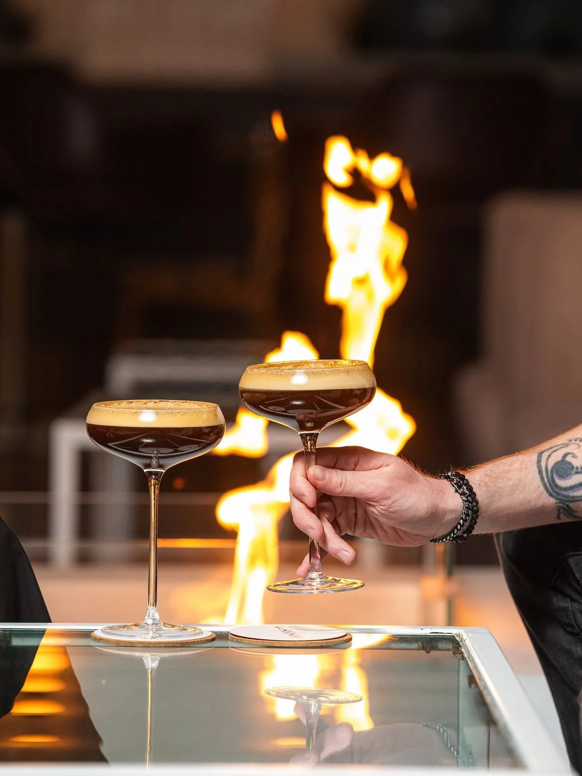 Two espresso martinis in elegant coupe glasses at Jensens in Kareela, featuring rich coffee foam, a glass tabletop and dramatic flames illuminating the background.