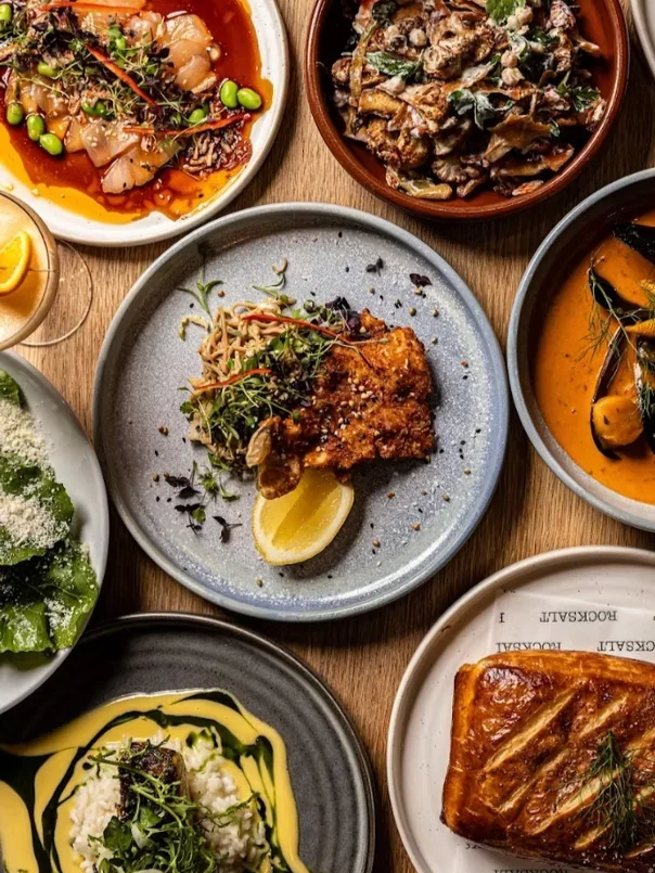 An overhead spread of shared plates, including sashimi, a seafood curry and a savoury pastry, on branded Rocksalt tableware.