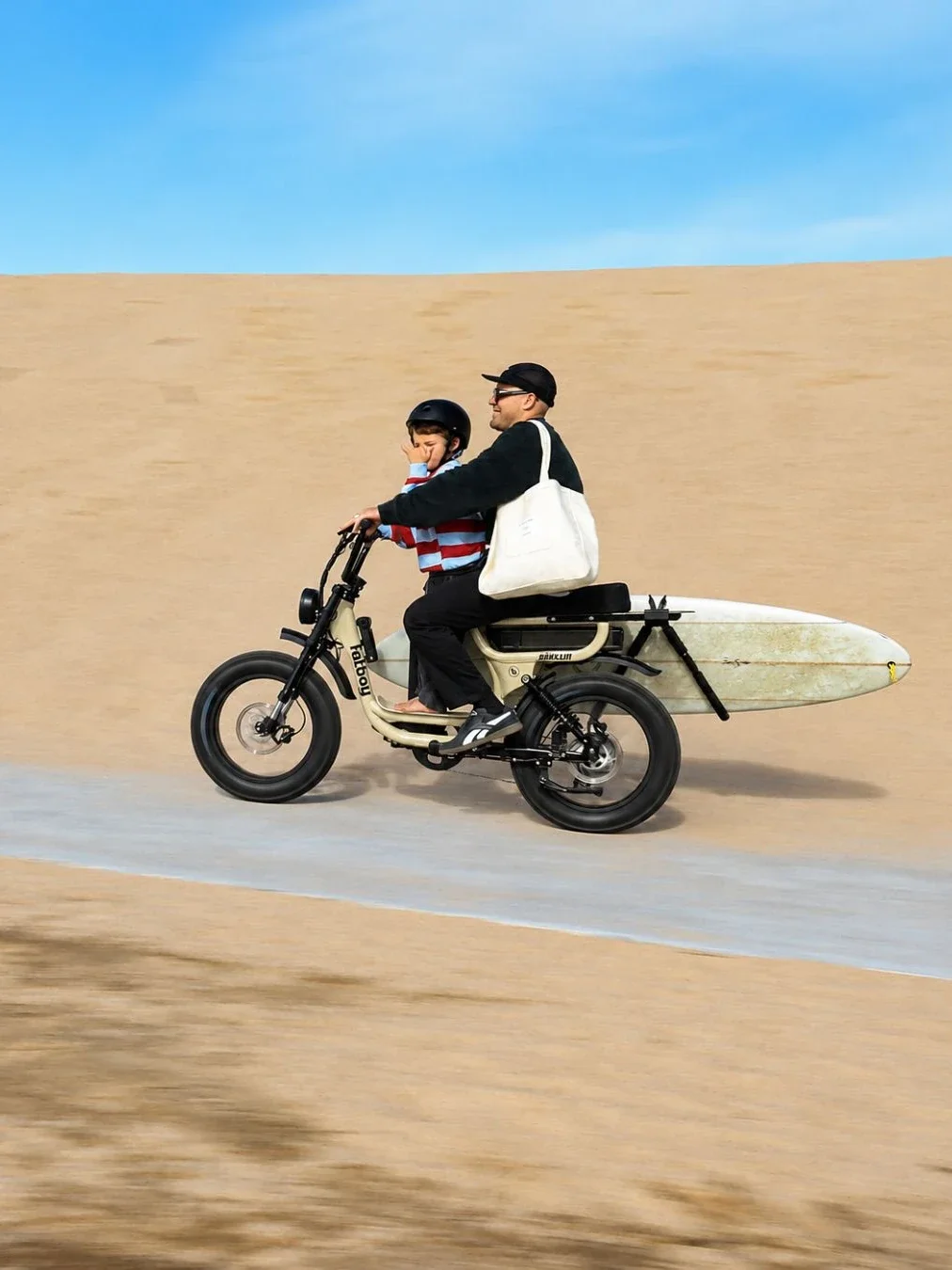A man and a young boy in a helmet riding a Fat Boy Bikes electric cargo bike along a beachfront path in Caringbah, with a white surfboard strapped to the rear rack and sand dunes behind.